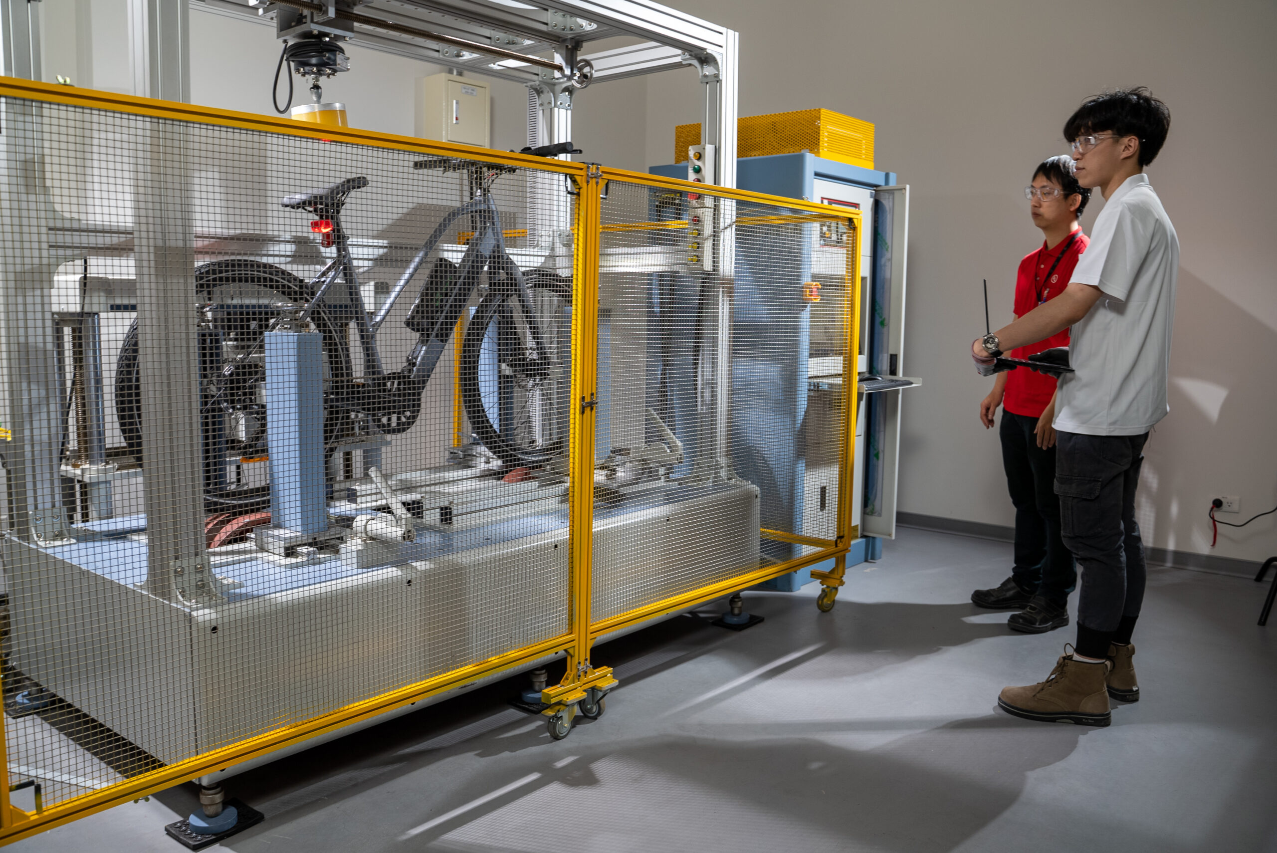 Two people observe a bicycle inside a safety enclosure as it undergoes mechanical testing by automated equipment in a laboratory setting.