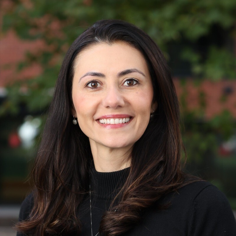 A woman with long brown hair and brown eyes smiles at the camera. She is wearing a black top and standing outdoors with green leaves and a brick building blurred in the background.