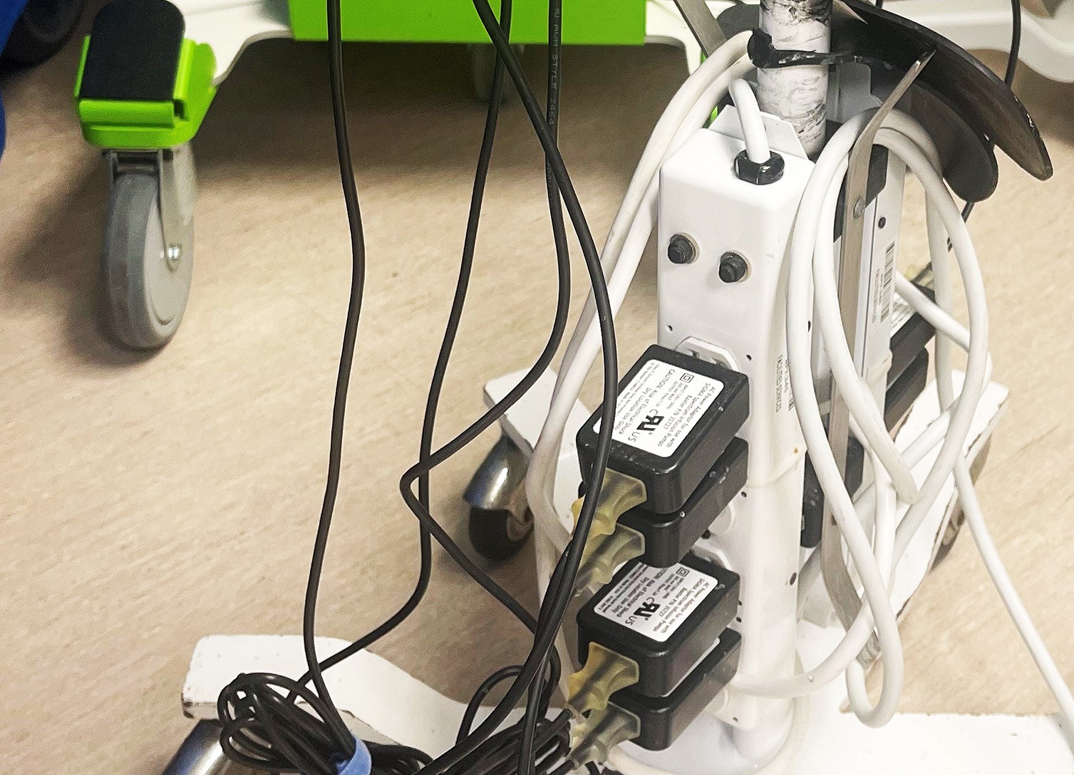 A power strip on the floor with multiple black and white cables and adapters plugged in, next to a green cart with wheels on a beige surface.