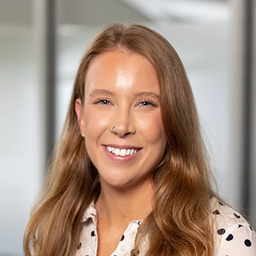 A woman with long, light brown hair smiles at the camera. She is wearing a white shirt with black polka dots and is in an office setting with blurred glass walls in the background.