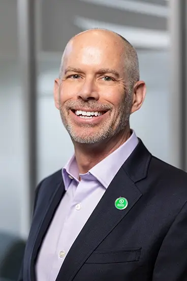A smiling, bald man with a trimmed beard wearing a dark blazer, lavender dress shirt, and a green circular pin, standing in front of a blurred office background.