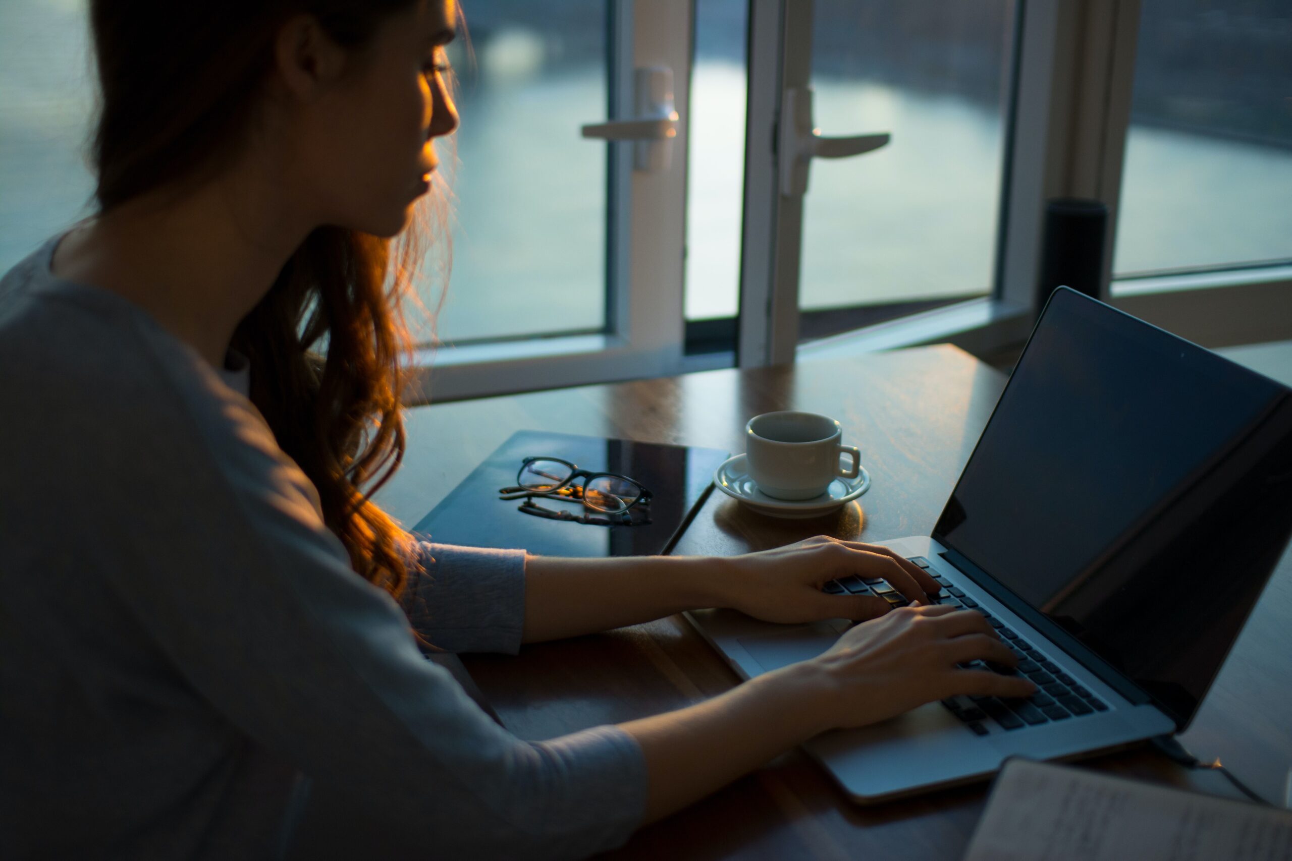 A person sits at a desk typing on a laptop, with a cup of coffee, a notebook, and glasses nearby. Soft light streams in through large windows in the background.