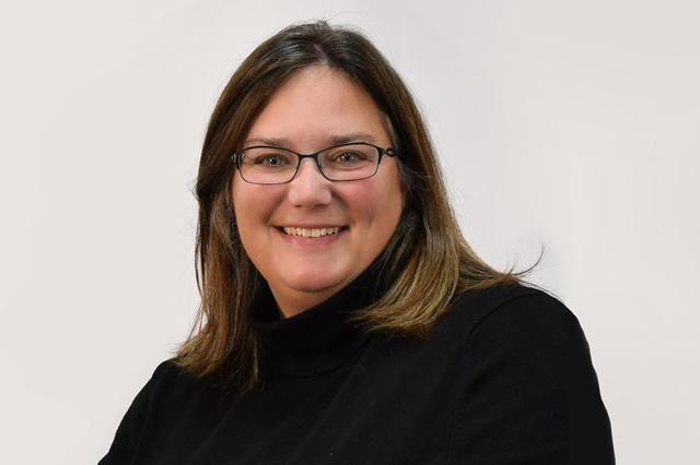 A woman with straight brown hair and glasses smiles at the camera. She is wearing a black turtleneck sweater, and the background is plain and light-colored.