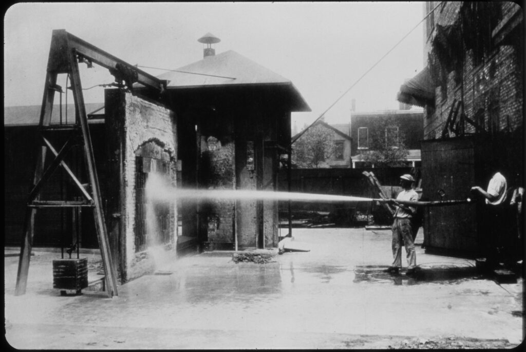 A person uses a fire hose to spray water at a brick structure outdoors, demonstrating the hose's strength. Buildings and trees are visible in the background.