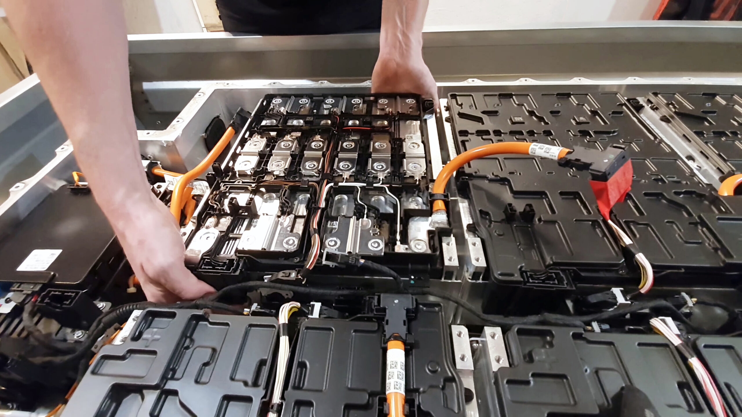 A person’s hands working on the internal components of an electric car battery pack, with visible orange cables and several interconnected battery cells.