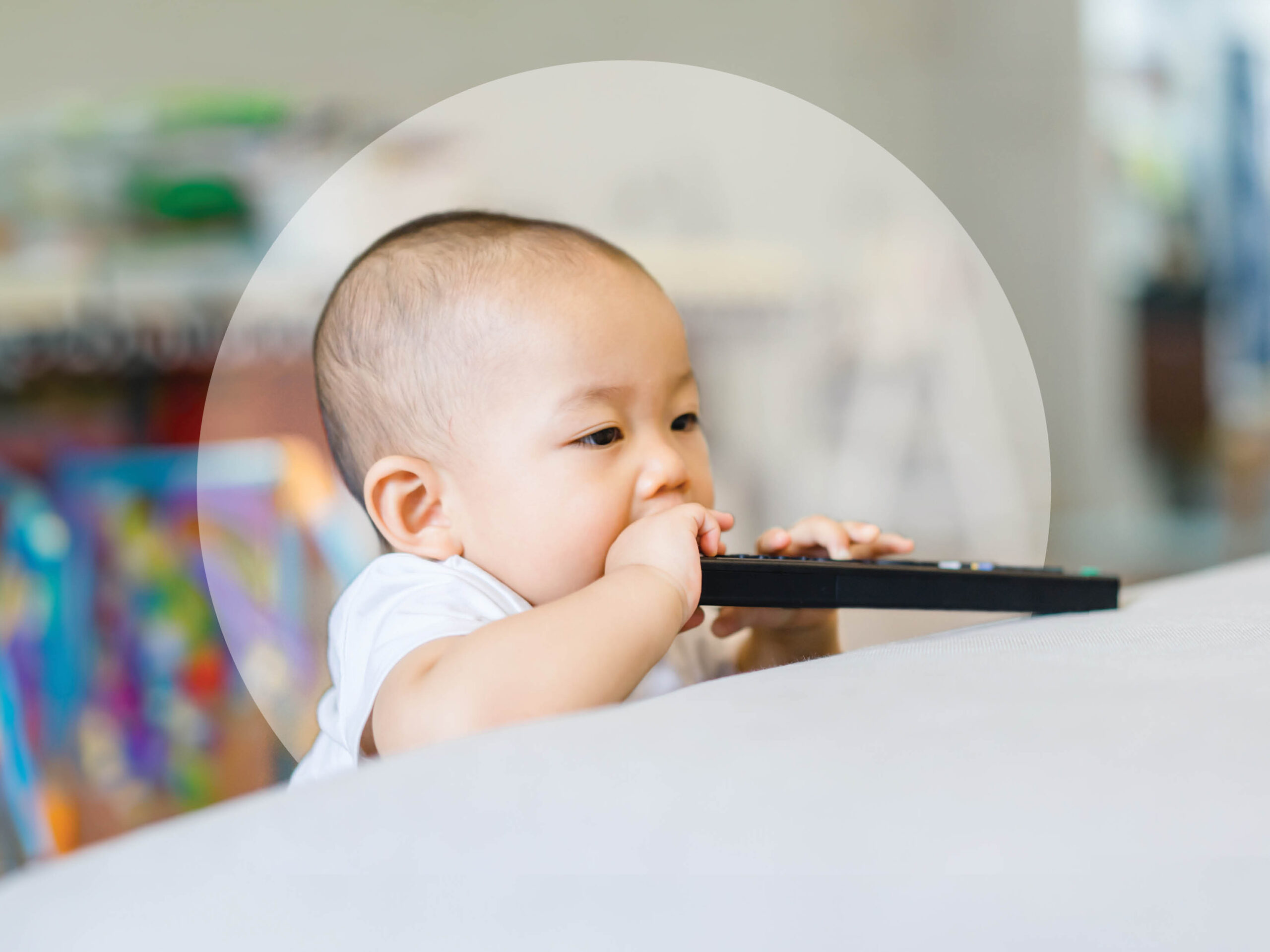 A baby stands against a white surface, holding and biting a television remote control. The background is softly blurred.