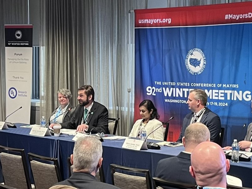 Five people sit at a panel table speaking at the 92nd U.S. Conference of Mayors Winter Meeting in Washington, D.C. Banners and microphones are visible, with attendees seated in the foreground.