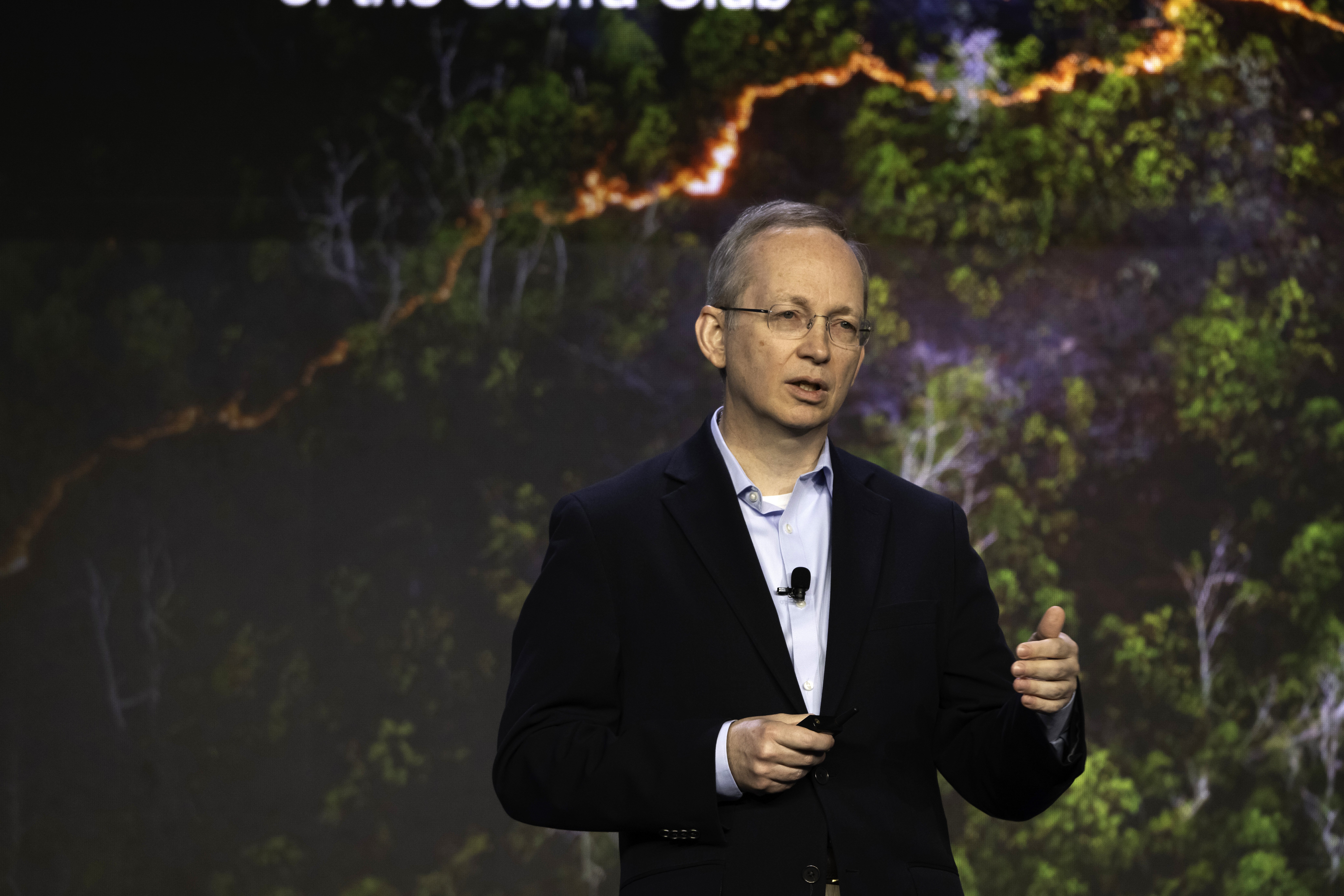A man in a dark suit and glasses speaks on stage, holding a clicker. Behind him is a large screen displaying an aerial view of a forest with a glowing, winding line through the trees.