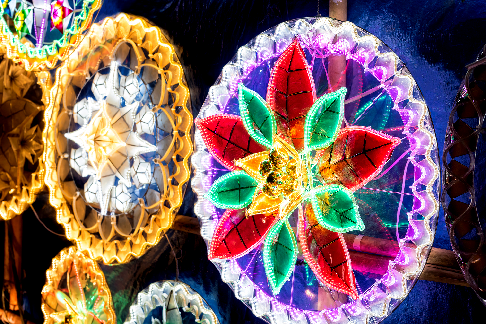 Colorful Filipino Christmas lanterns, known as parols, are lit up at night. The lanterns have intricate patterns, with bright red, green, yellow, and white lights arranged in star and flower shapes.