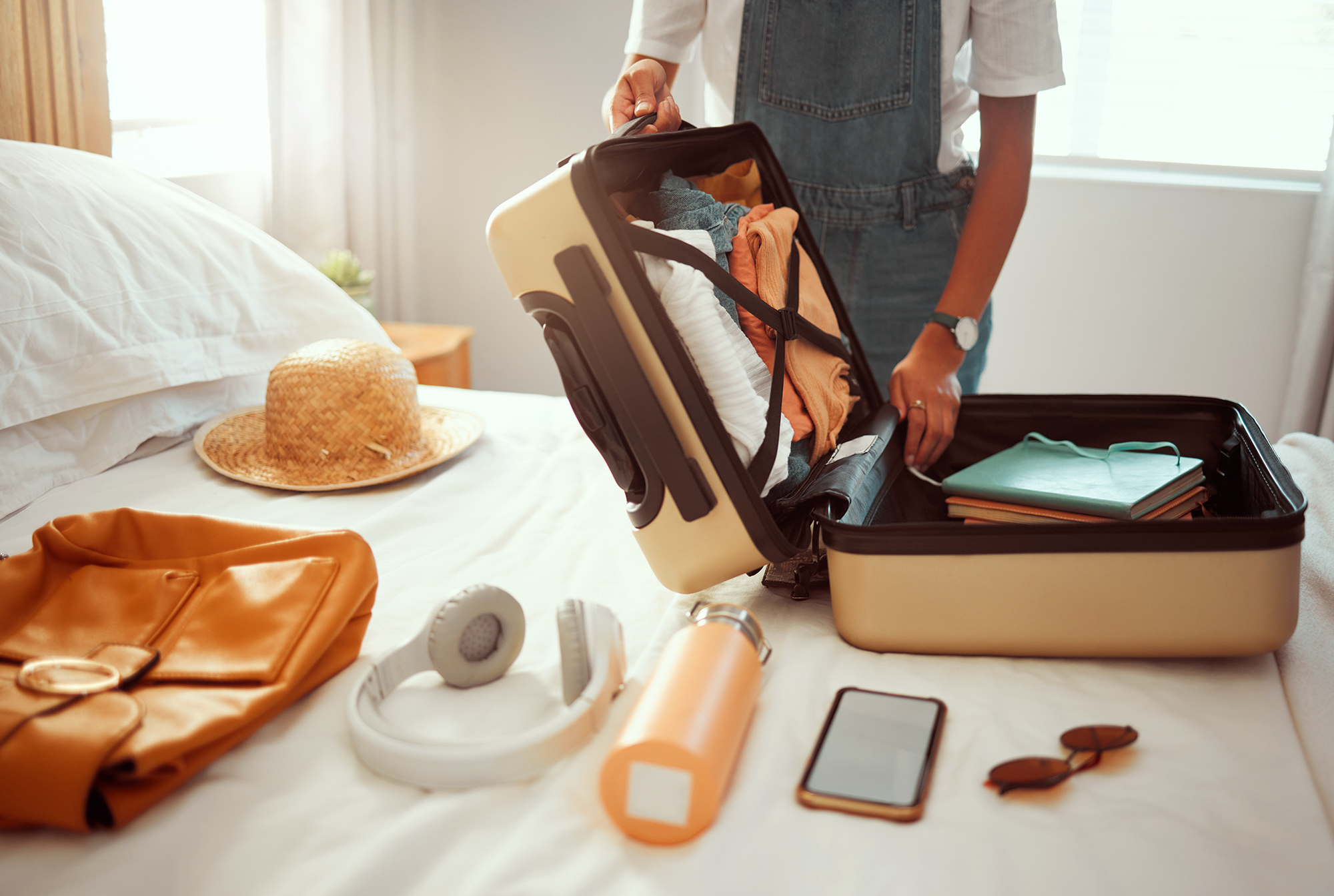 A person packing a beige suitcase on a bed with clothes, a hat, headphones, a water bottle, a smartphone, sunglasses, and a jacket laid out nearby. Sunlight streams through a window in the background.