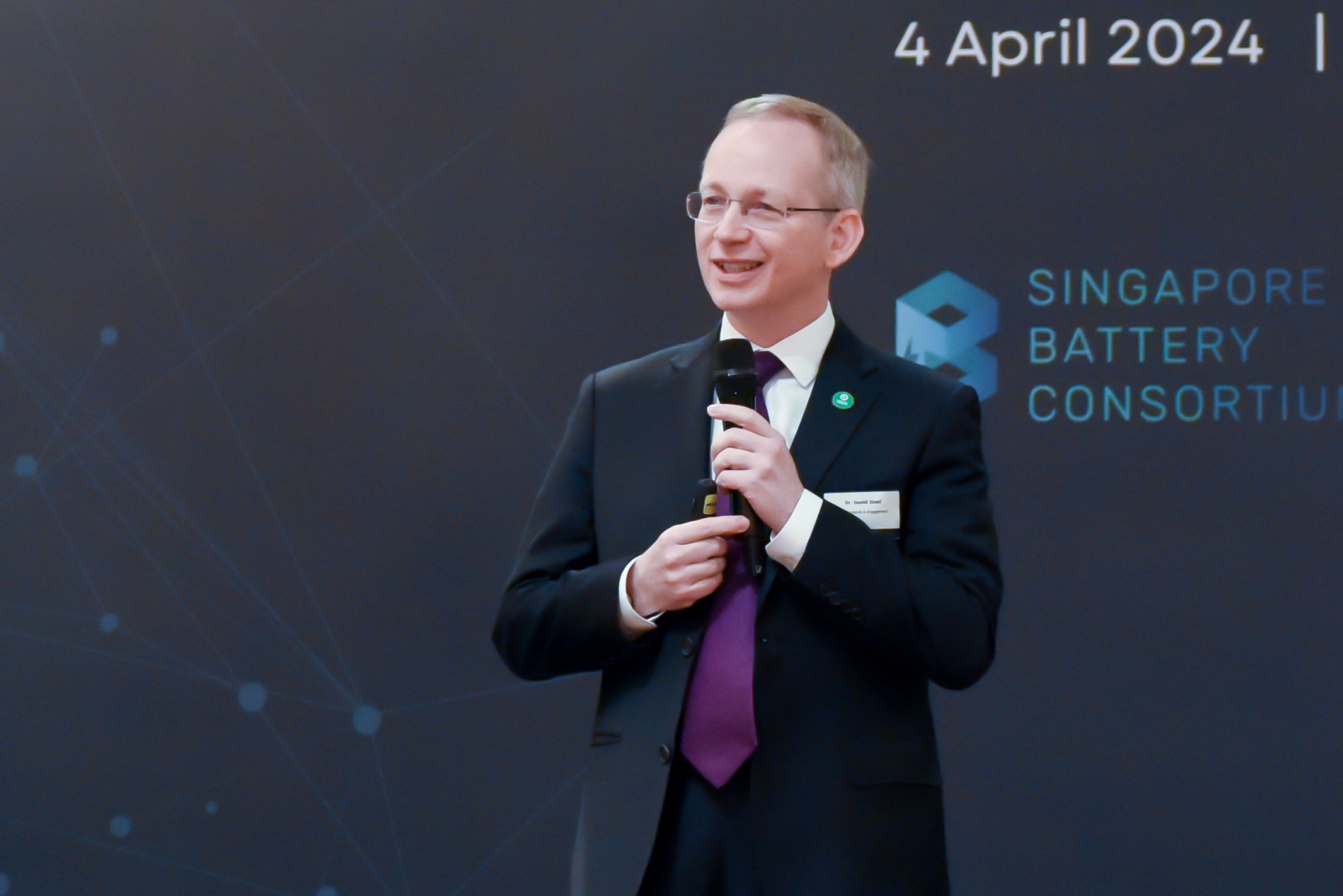 A man in a suit and purple tie holds a microphone and speaks at an event for the Singapore Battery Consortium on 4 April 2024, standing in front of a dark backdrop with event branding.