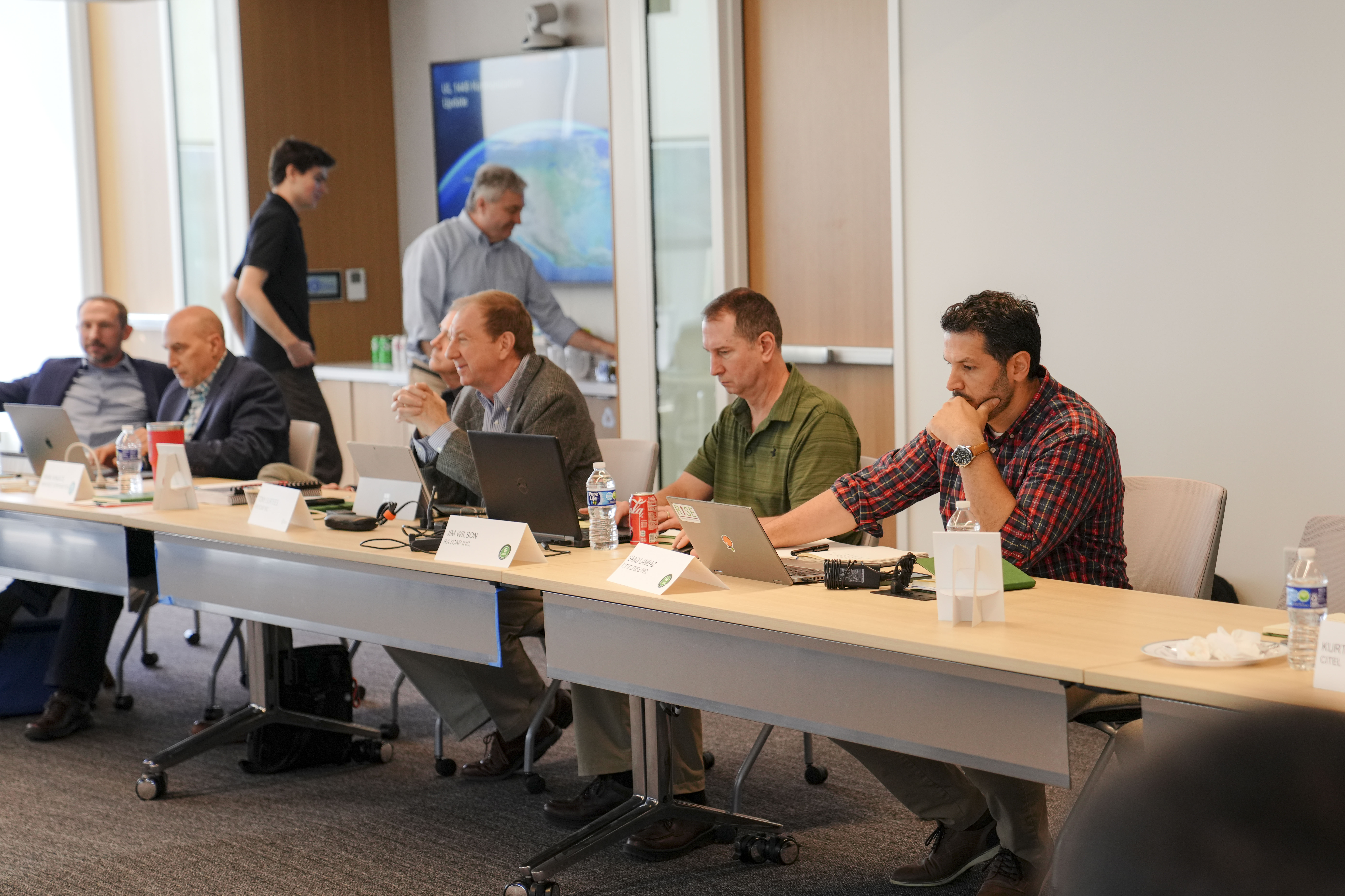 A group of men sit at a long conference table with laptops and name cards, focused on a meeting. One man stands in the background near a table with snacks and drinks. The room is well-lit and modern.