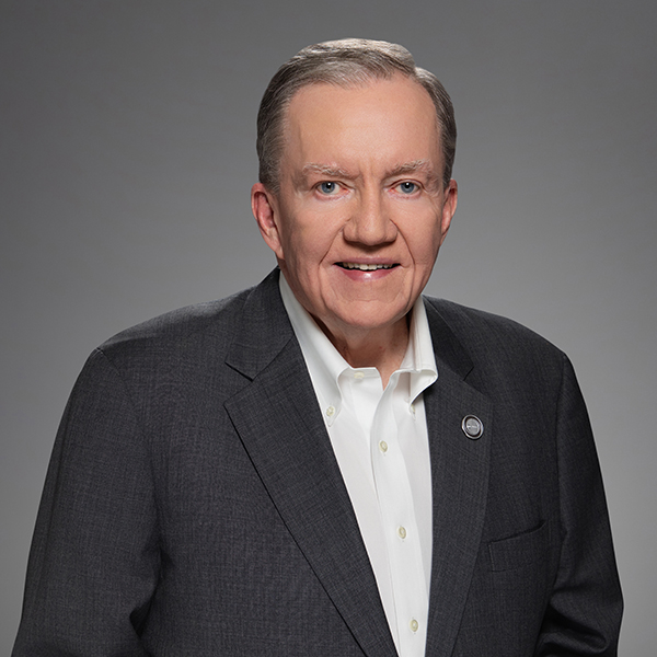 An older man with short gray hair smiles, wearing a dark suit jacket over a white button-down shirt, standing against a plain gray background.
