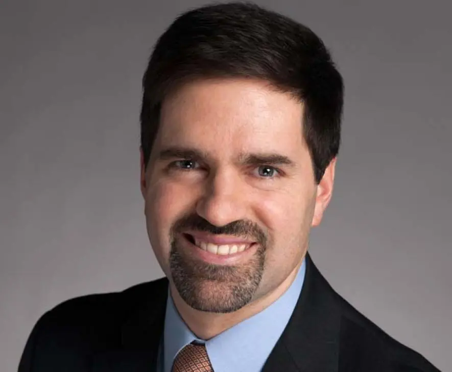 A man with short dark hair, a trimmed beard and mustache, wearing a dark suit, light blue shirt, and patterned tie, smiling at the camera against a plain gray background.