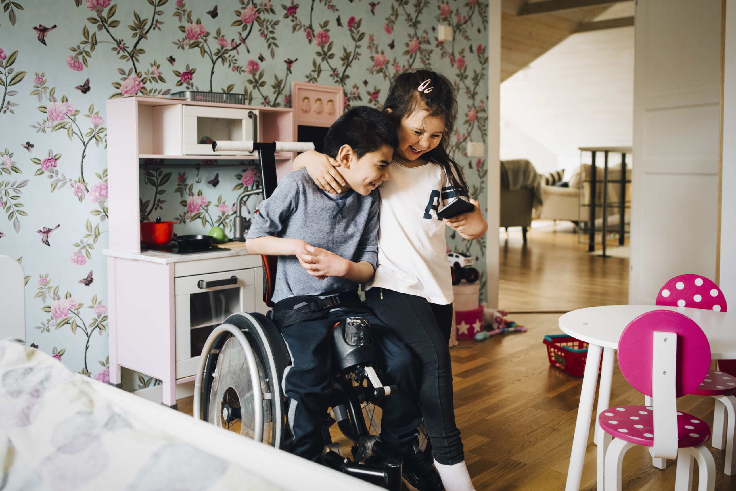 Two children smile and hug in a playroom; one uses a wheelchair, and the other holds a phone. They stand near a toy kitchen set against a floral wallpaper background, with a small table and chairs nearby.