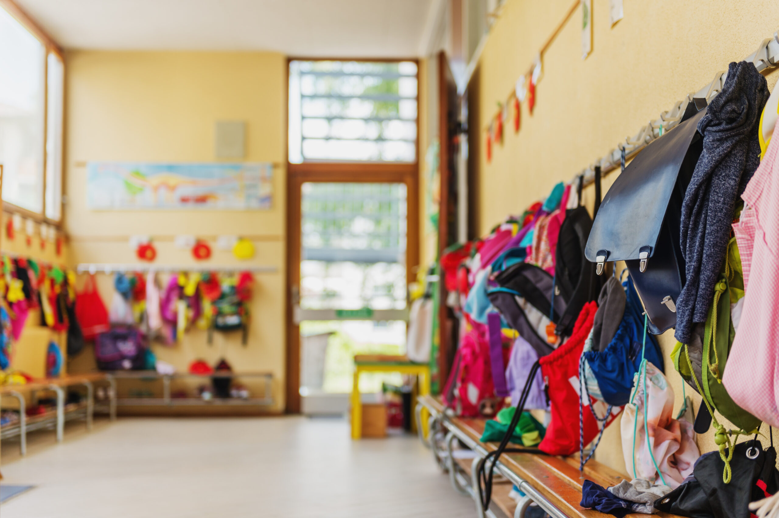 A bright, colorful school hallway with backpacks, jackets, and bags hanging on hooks along the wall. Benches are placed below, and sunlight streams in through large windows and an open door at the end of the hall.