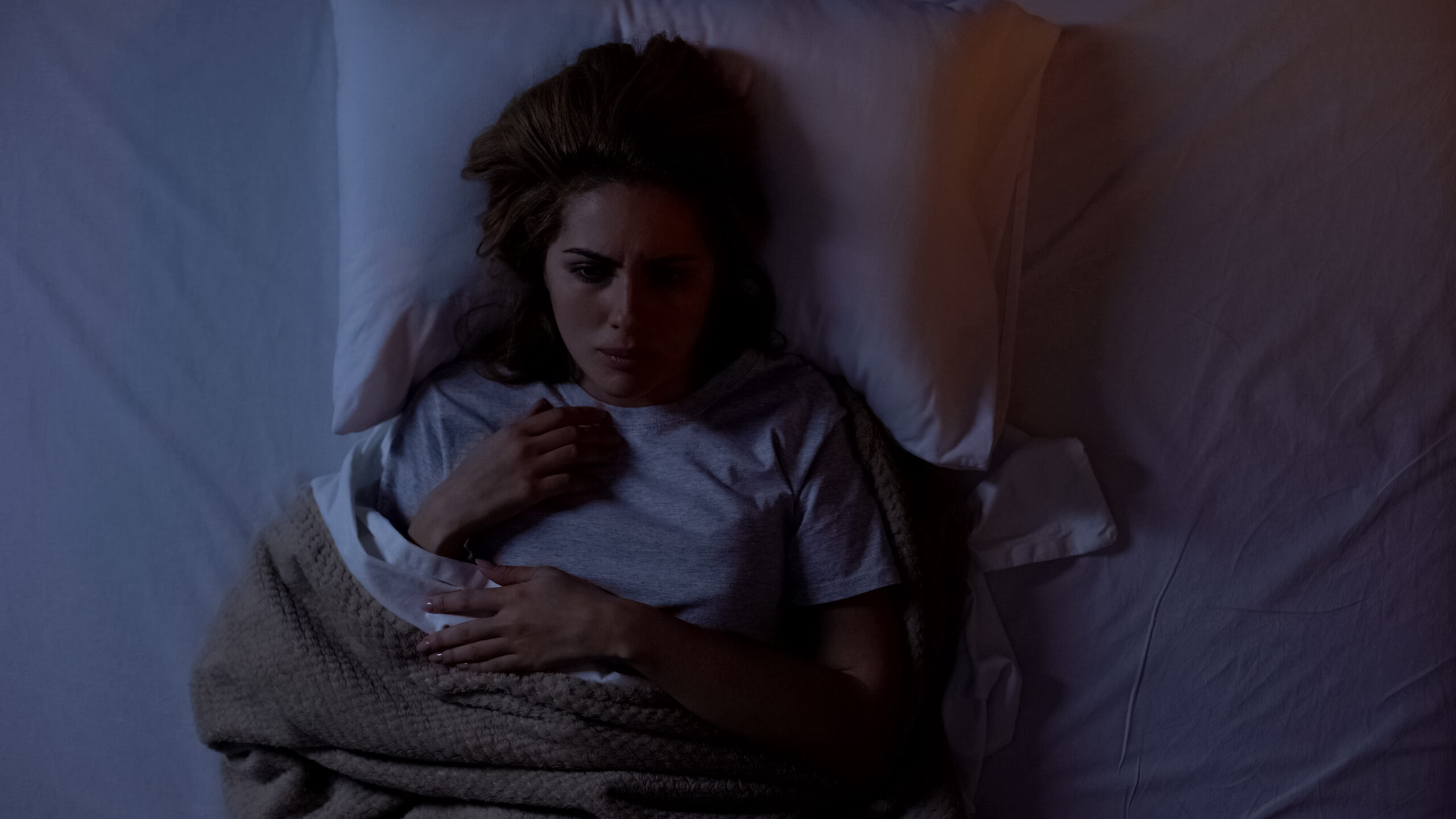 A woman lies in bed at night under a blanket, looking anxious and unable to sleep, with one hand on her chest and a worried expression on her face. The lighting is dim and somber.