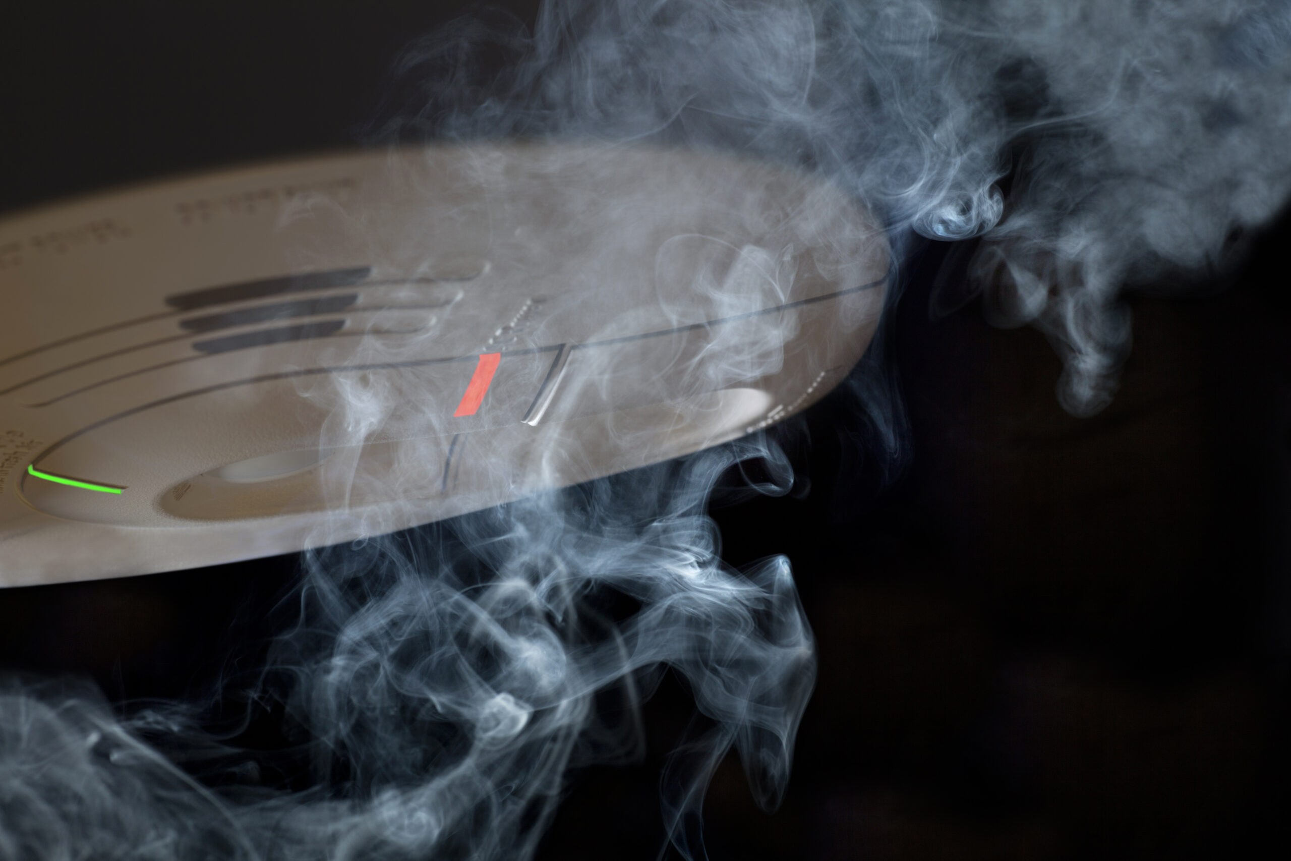 A close-up of a smoke detector surrounded by swirling smoke, with the device's indicator lights clearly visible.