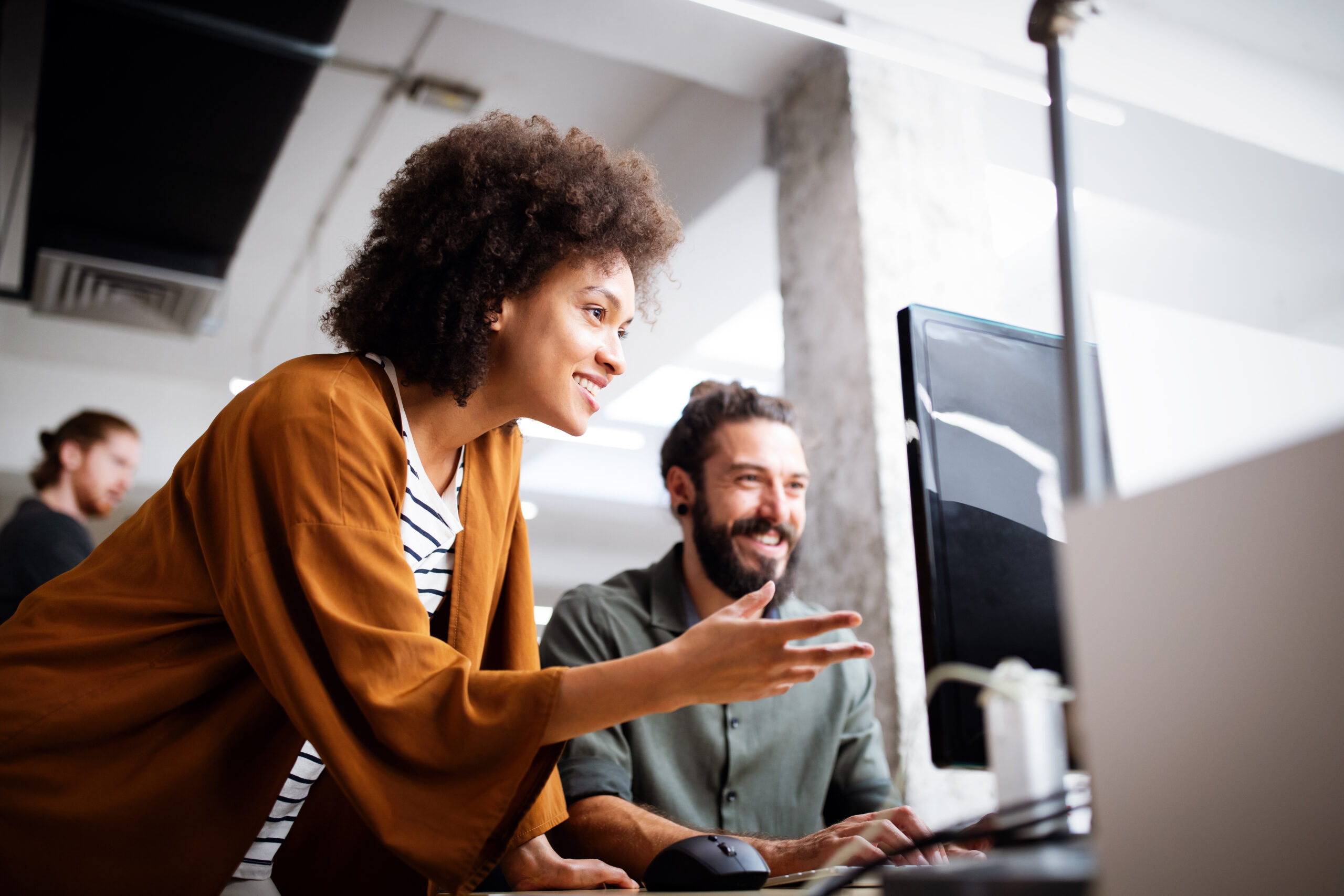 Two people smiling and looking at a computer monitor in a bright, modern office. One person is standing and gesturing toward the screen while the other is seated, both appearing engaged and collaborative.