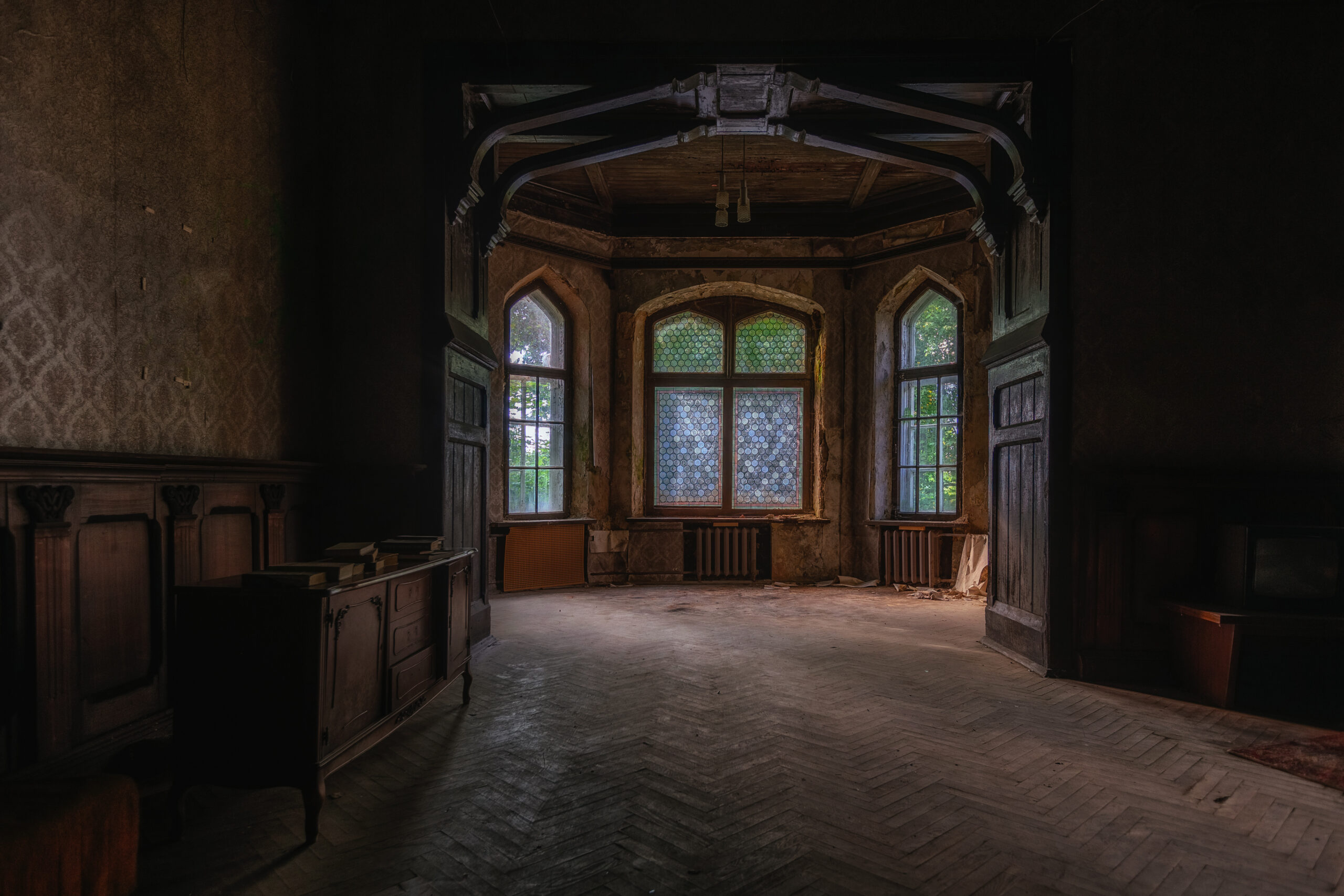 A dimly lit, abandoned room with ornate wooden panels, arched windows with stained glass, and dusty, worn floors. Natural light filters through the windows, highlighting the room’s faded, eerie elegance.