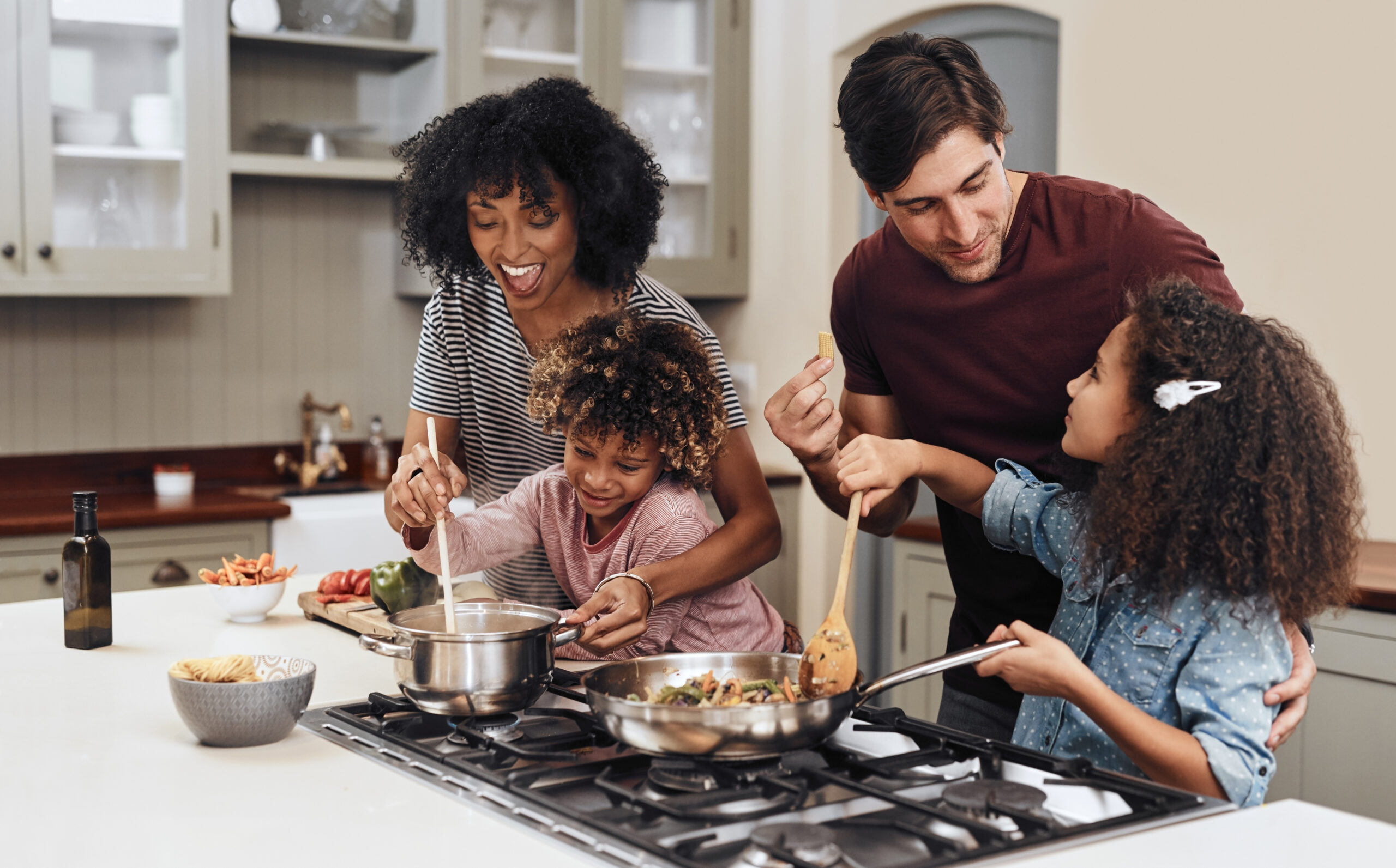 A happy family of four cooks together in a modern kitchen, smiling and stirring food in pans on the stove. The parents and two young children, one holding a spoon, enjoy the activity and each other’s company.