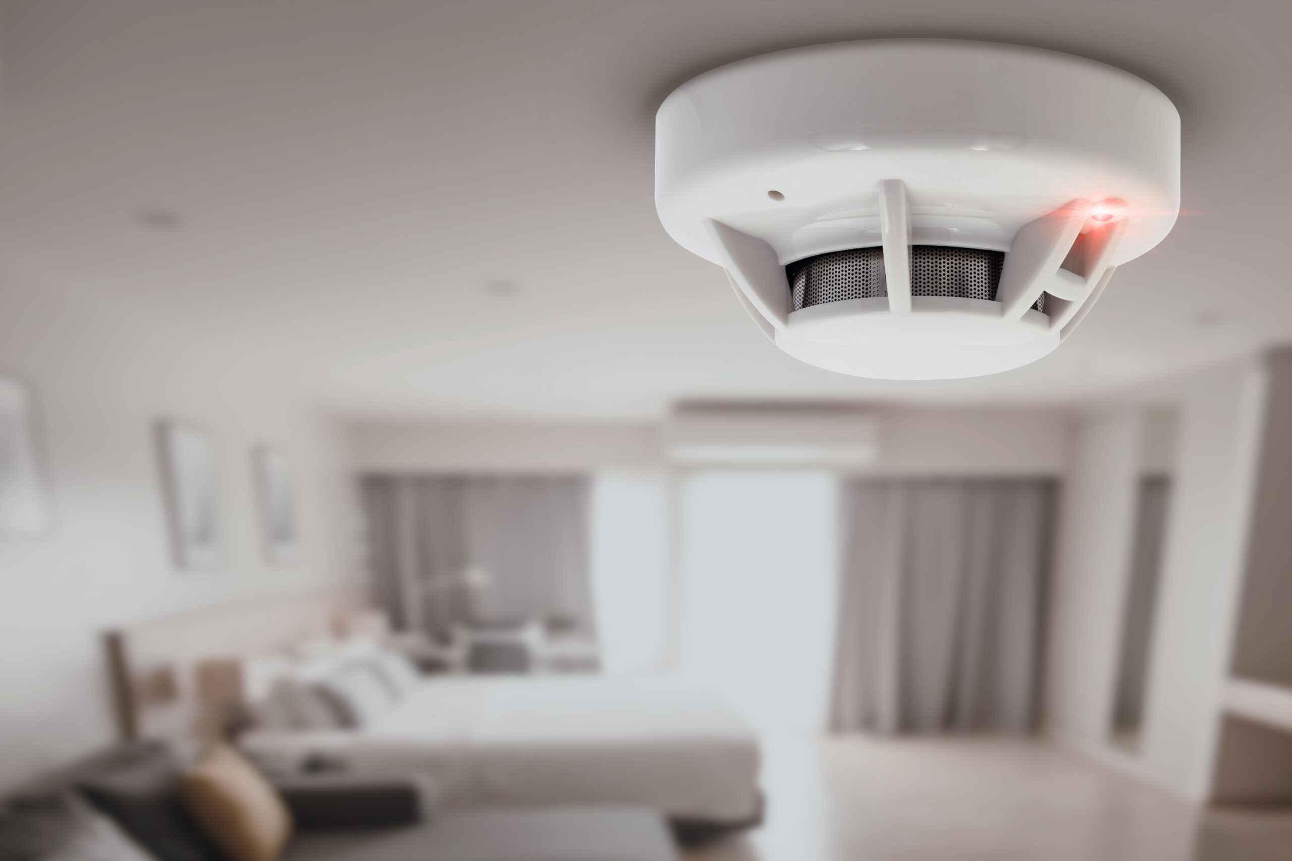 A smoke detector with a red indicator light is mounted on the ceiling of a modern, softly lit bedroom with a bed, cushions, and large windows in the background.