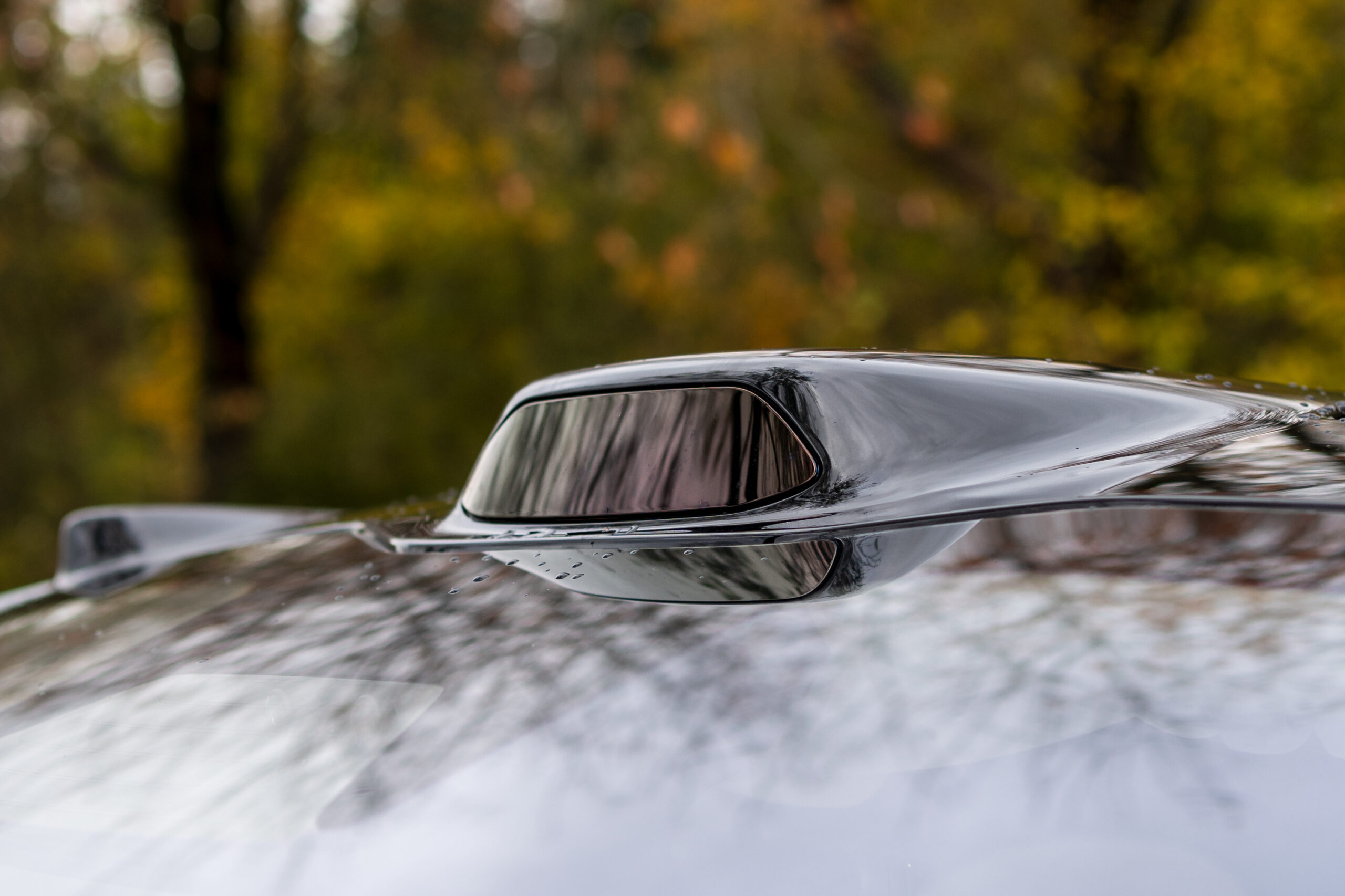 A close-up of a black car roof shows a smooth, aerodynamic shark fin antenna with blurred autumn trees in the background.