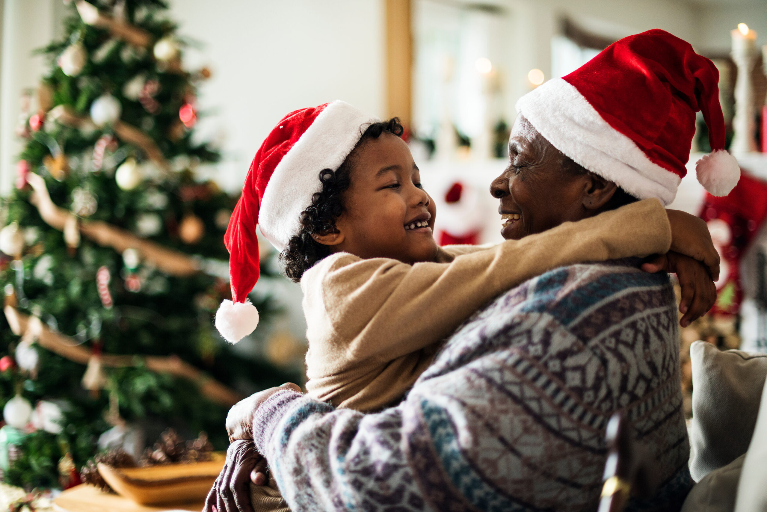 A child and an older adult wearing Santa hats smile and hug each other in front of a decorated Christmas tree at home.