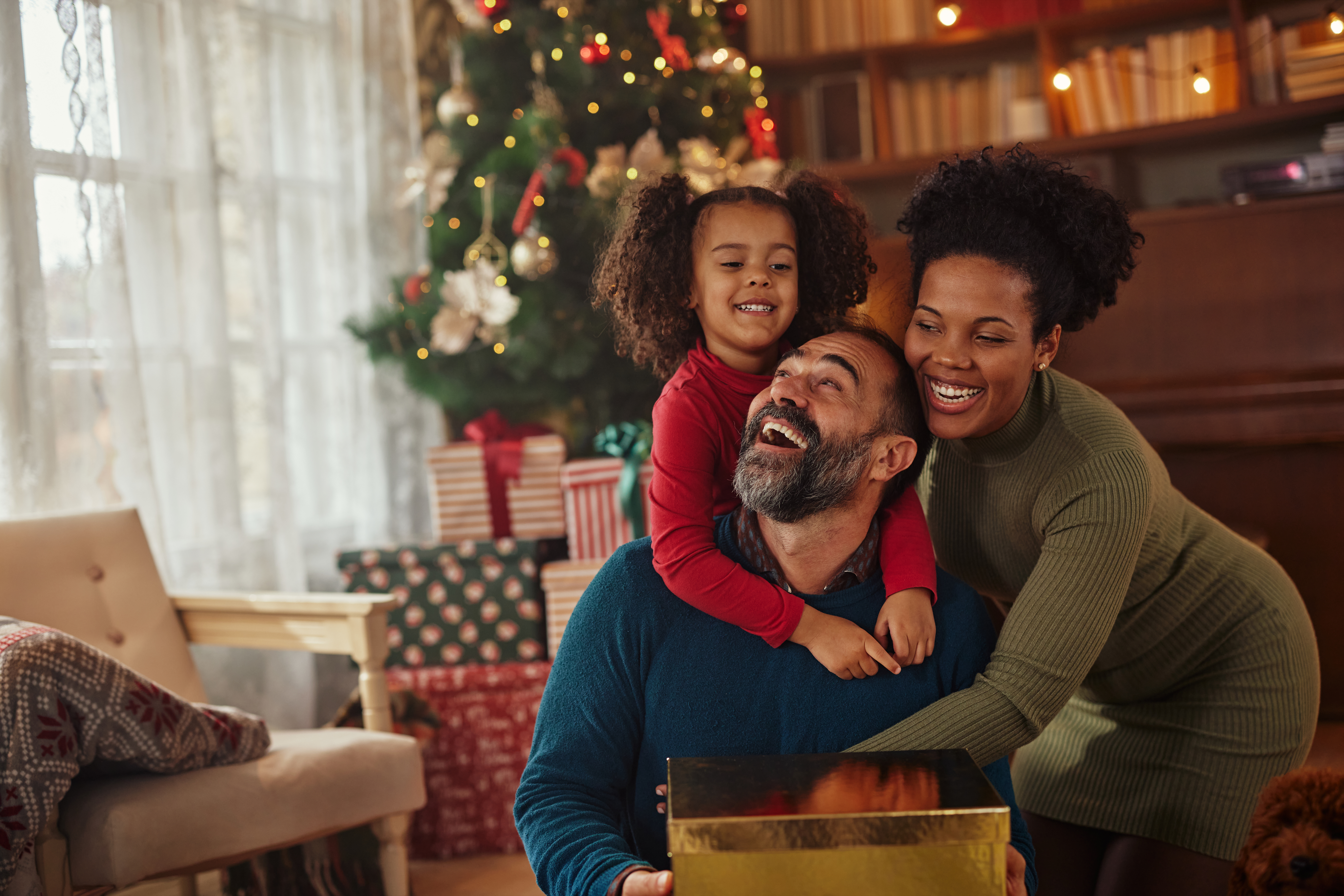 A smiling family sits together by a Christmas tree, surrounded by wrapped gifts. A child in red hugs an adult man, while an adult woman leans in close, all looking happy and festive. Warm lights and decor complete the scene.