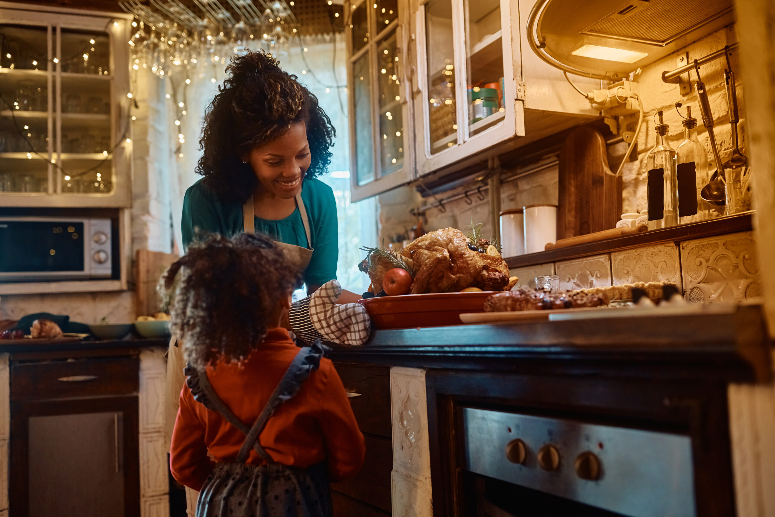A woman in an apron smiles at a child in a cozy, warmly lit kitchen decorated with string lights. A roasted turkey and side dishes are on the counter, suggesting a festive meal preparation.