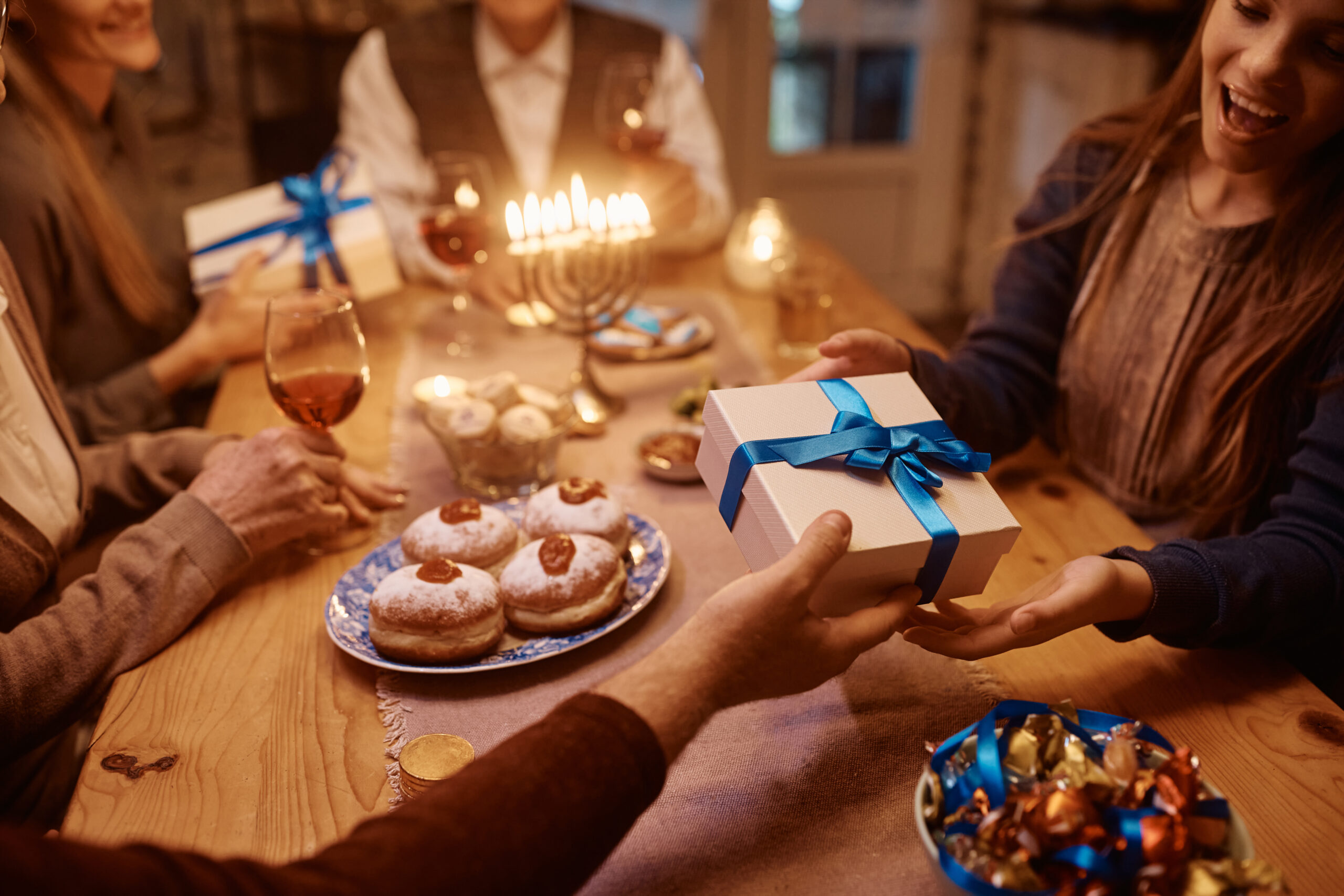Four people sit at a festive table with candles, donuts, and chocolates. One person hands a blue-ribboned gift box to a smiling woman, creating a warm, celebratory atmosphere.