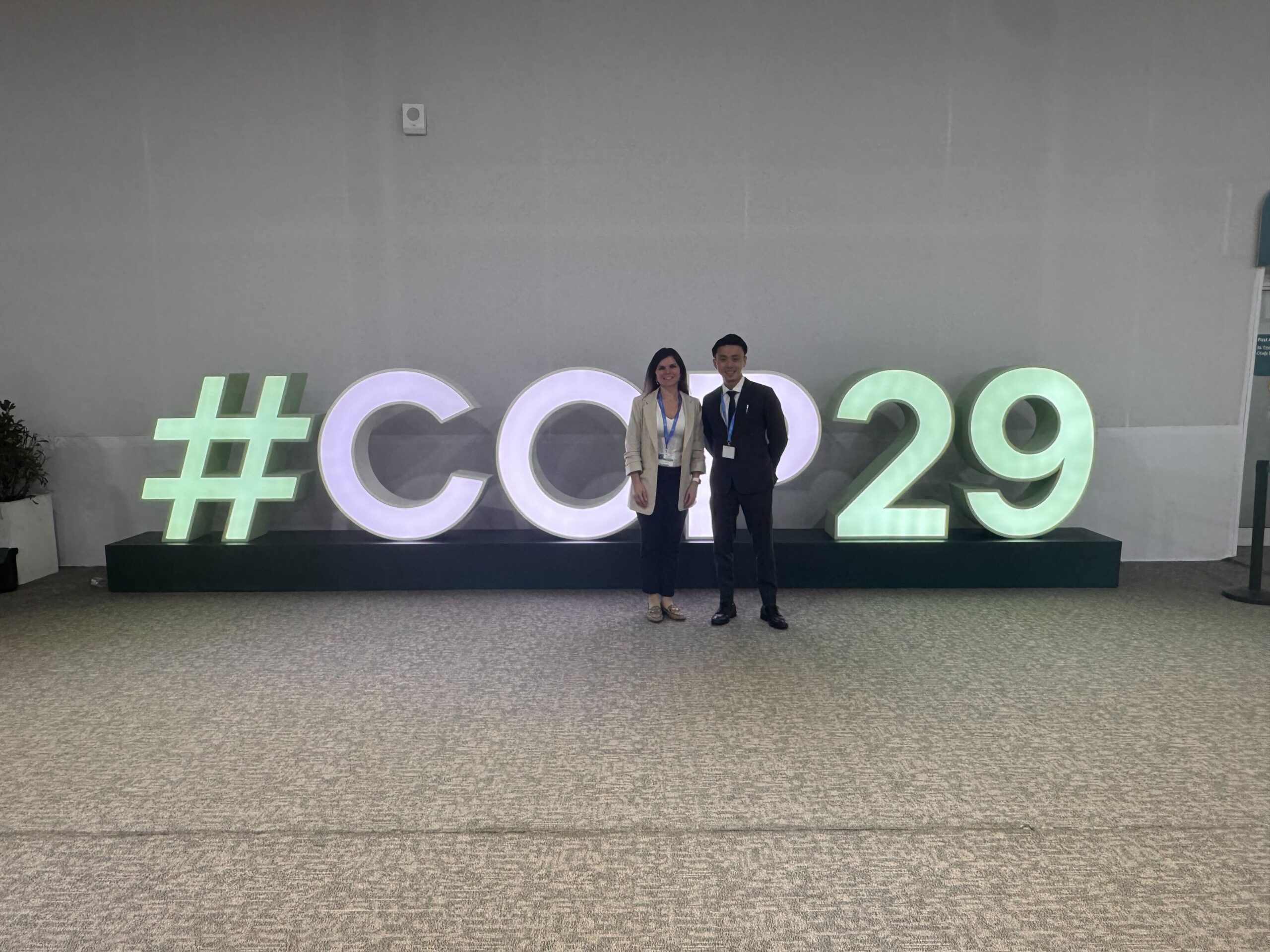 Two people stand in front of a large illuminated "#COP29" sign, posing for a photo in a spacious indoor venue with a light grey wall and carpeted floor.