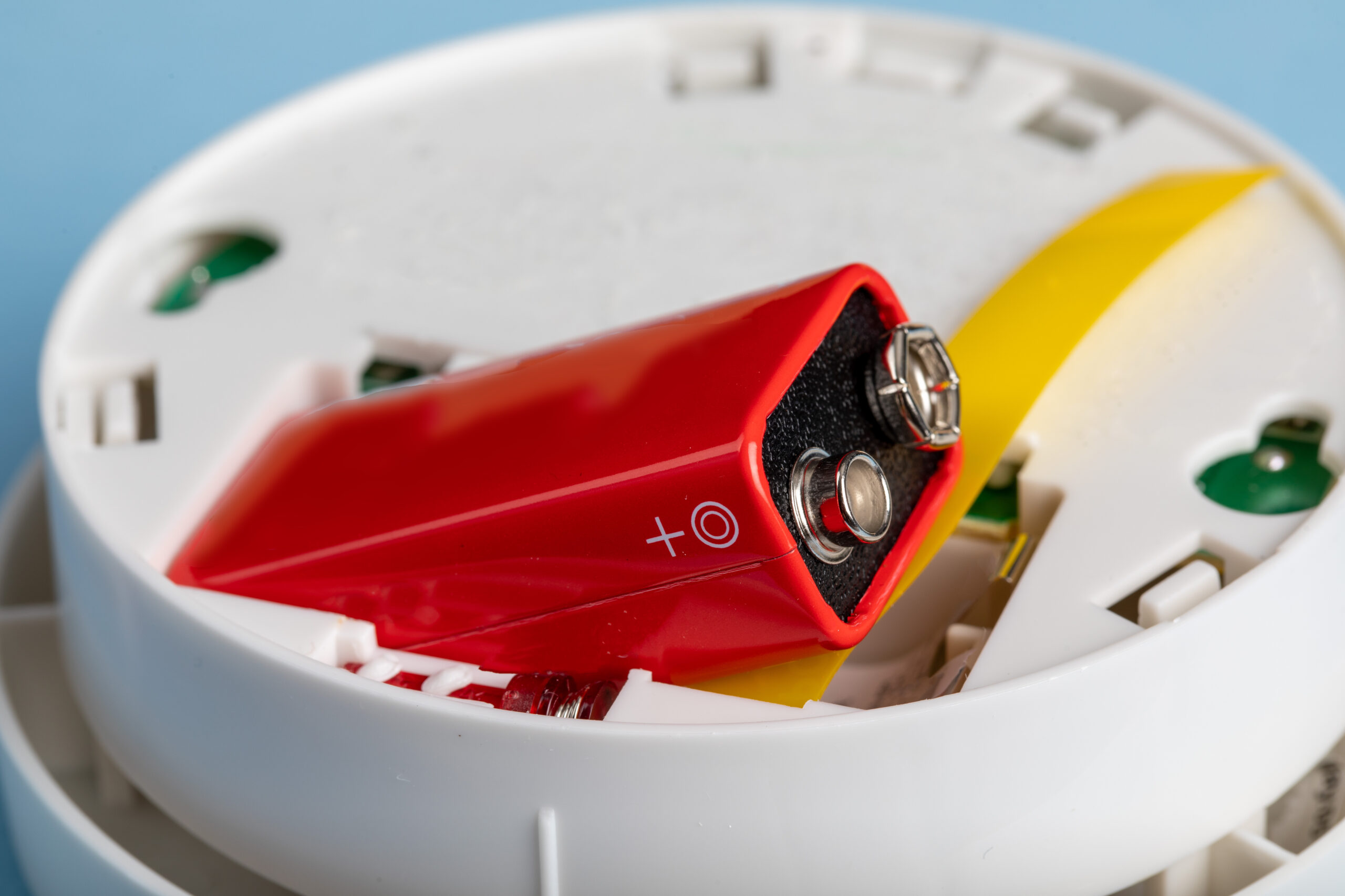 A close-up of a red 9-volt battery partially inserted into a white smoke detector with the cover open, showing internal components and a yellow ribbon inside.