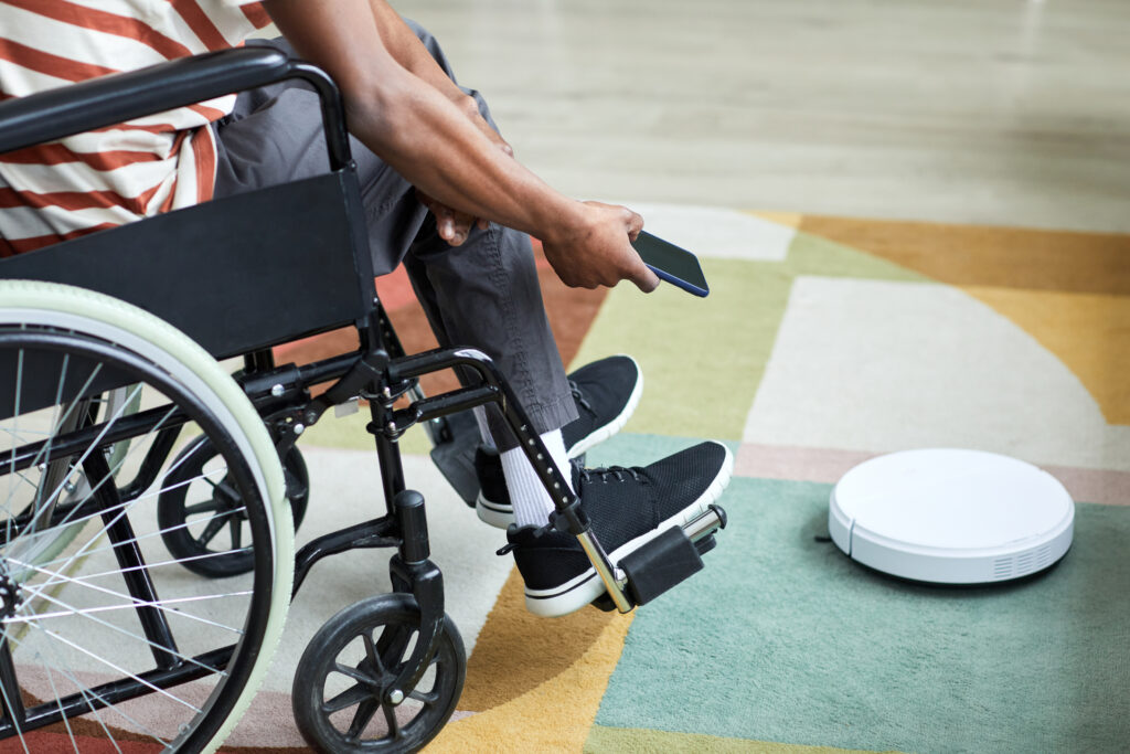 A person in a wheelchair uses a smartphone to control a white robotic vacuum cleaner on a colorful rug indoors.