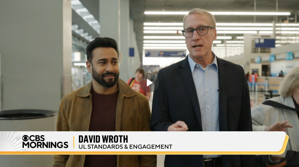 Two men stand and talk inside an airport terminal. The left man has dark hair and a beard, wearing a brown jacket. The right man wears glasses and a blue shirt. Text reads: "CBS Mornings, David Wroth, UL Standards & Engagement.