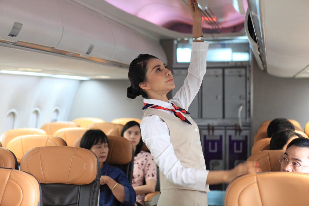 A flight attendant in uniform reaches up to an overhead bin inside an airplane cabin while passengers are seated in tan leather seats.