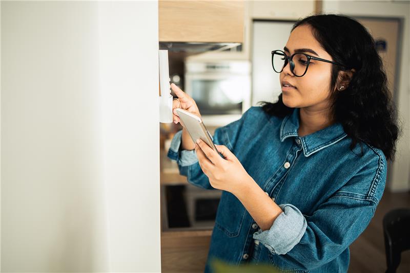 A person wearing glasses and a denim shirt uses a smartphone while adjusting a wall-mounted smart home device in a modern kitchen.