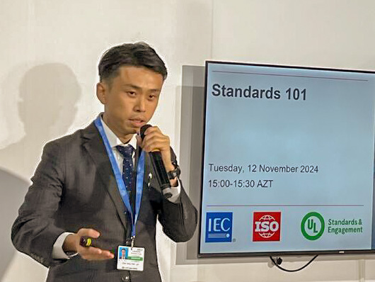 A man in a suit speaks into a microphone while pointing, standing next to a screen displaying "Standards 101" and logos for IEC, ISO, and UL. The event is scheduled for November 12, 2024, at 15:00-15:30 AZT.