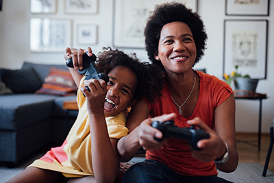 A smiling woman and young girl sit on a couch, enthusiastically playing video games together with controllers in hand, in a cozy living room decorated with framed pictures.