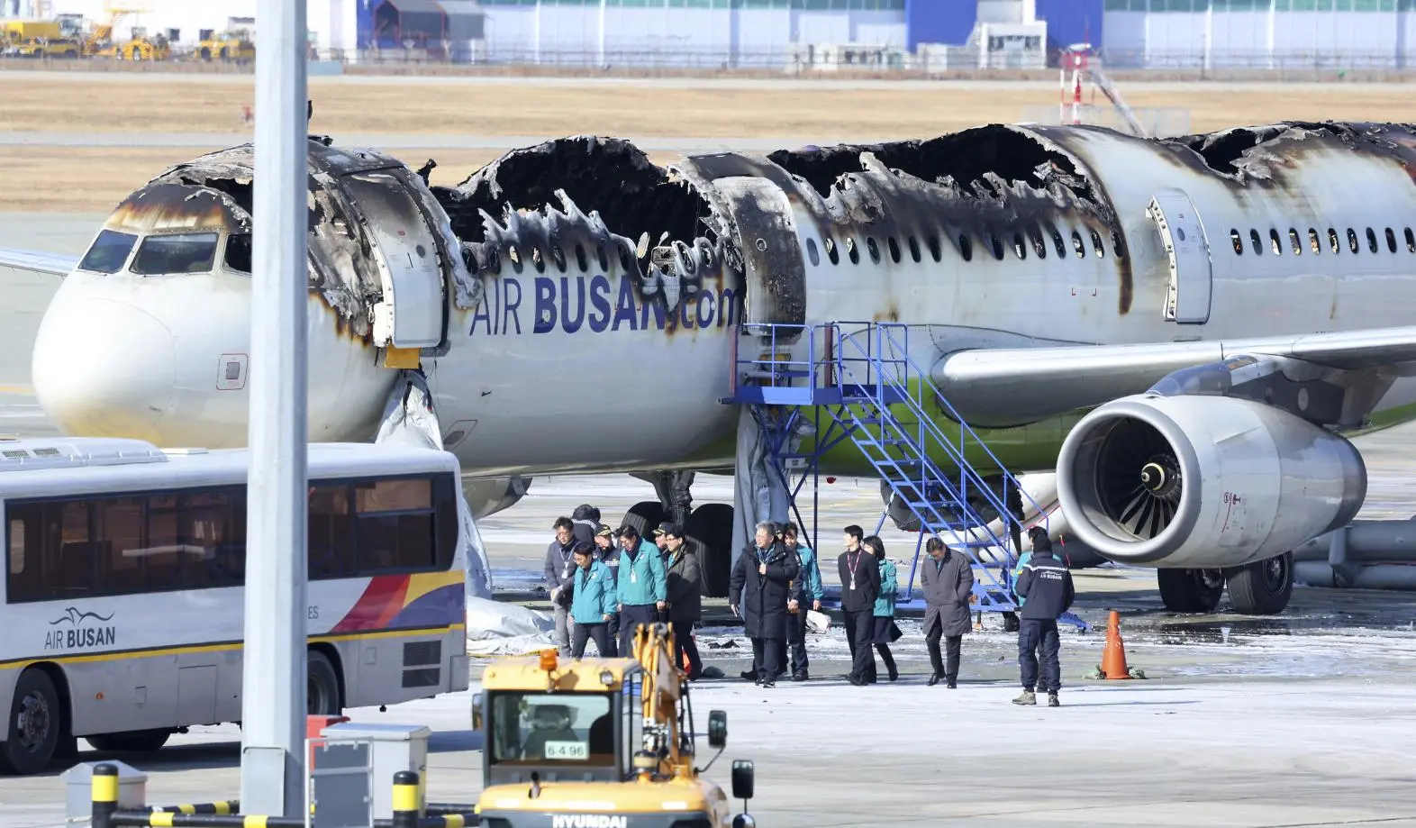 A large passenger airplane with severe fire damage along the top is parked on the tarmac. Several people in uniforms and safety vests stand near the aircraft, and airport vehicles are nearby.