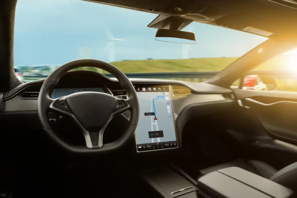 The interior of a modern car with a digital display showing navigation, a steering wheel, and empty seats, driving on a road with green fields and wind turbines visible outside at sunset.