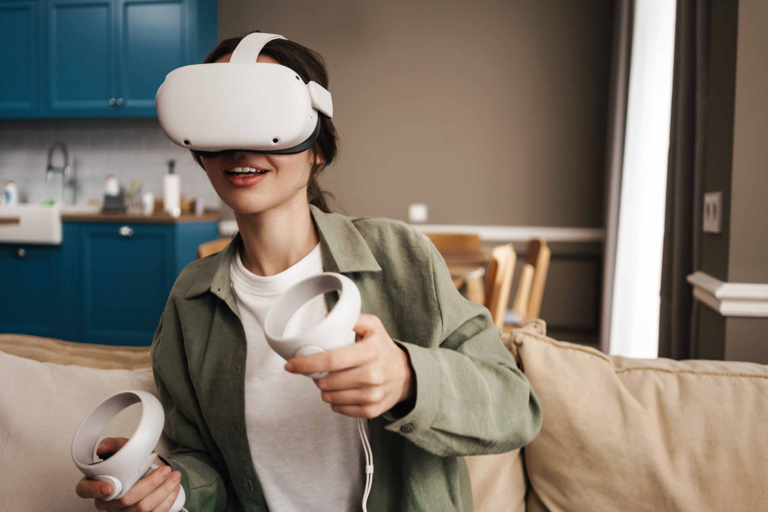 A woman sits on a beige couch wearing a virtual reality headset and holding two VR controllers, smiling and appearing engaged. A kitchen area with blue cabinets is visible in the background.