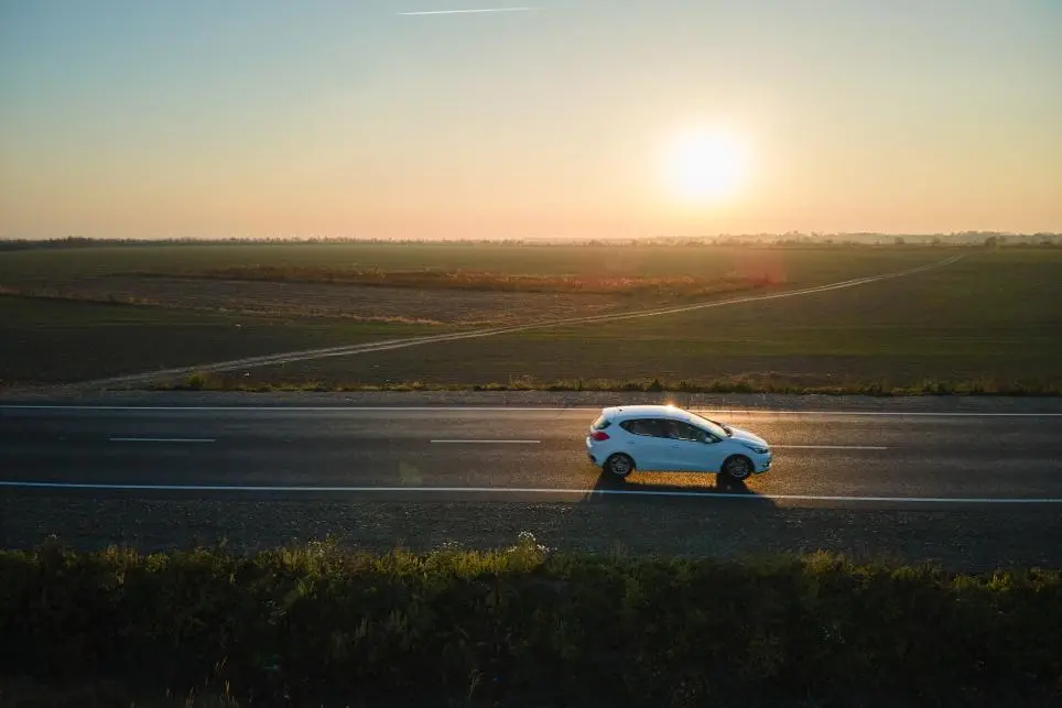 A white car drives on an empty road through flat farmland at sunset, with the sun low in the sky and fields stretching to the horizon.