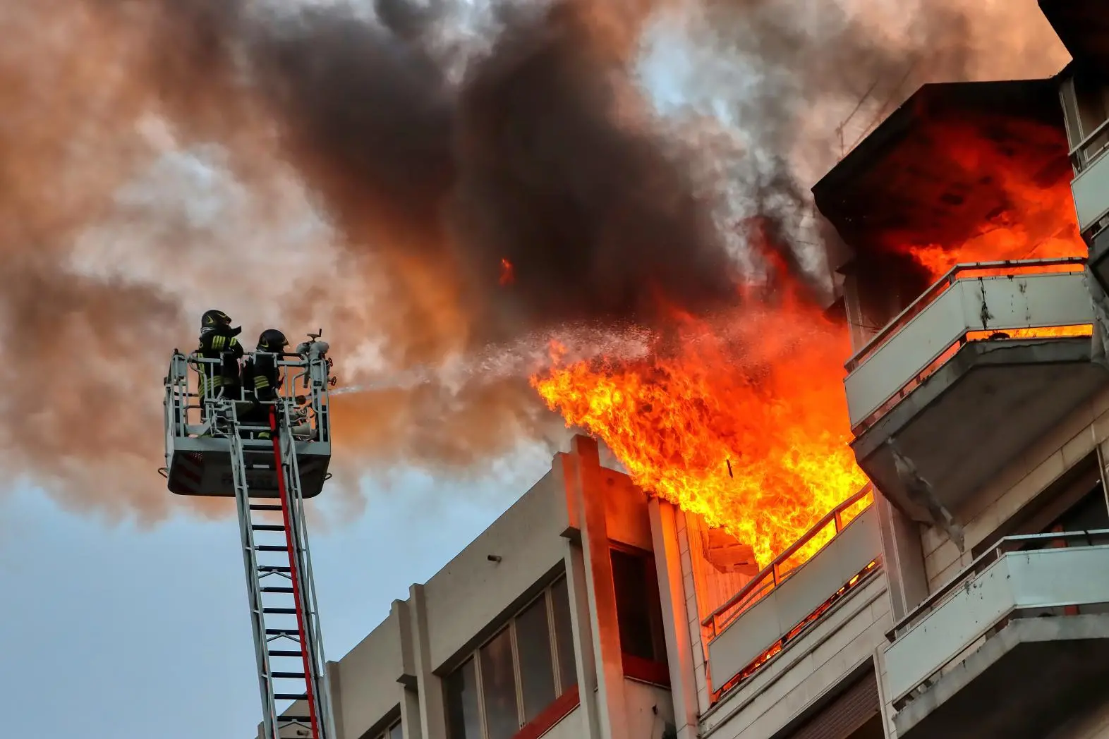 Two firefighters on an elevated platform spray water at intense flames and thick smoke pouring from the upper floors of a burning building, while nearby balconies appear empty and undamaged.