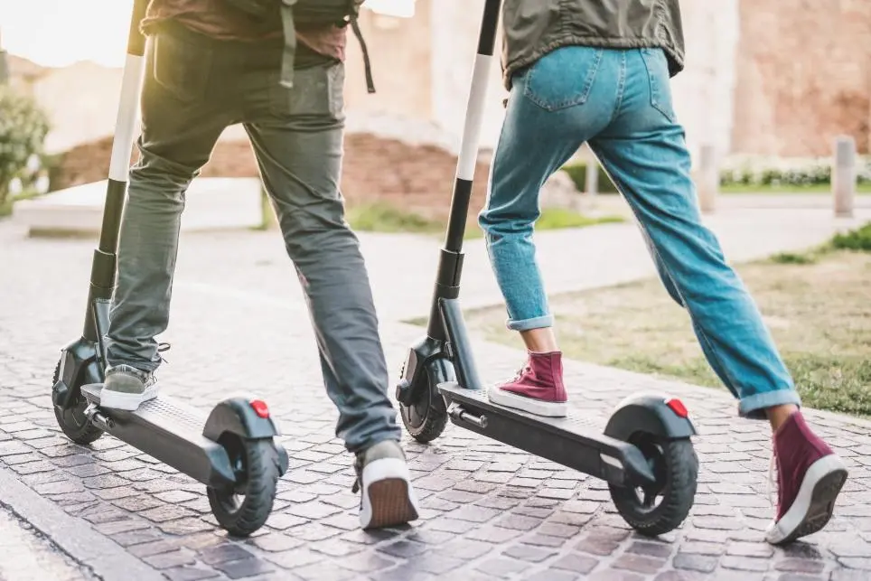 Two people ride electric scooters on a cobblestone path outdoors. Both wear casual clothes and sneakers, with only their lower bodies visible.