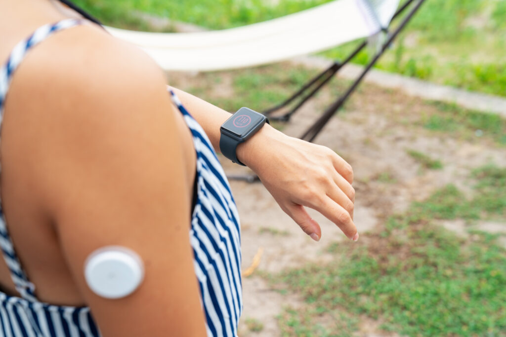 A person in a striped sleeveless top wearing a smartwatch checks their wrist. A round medical sensor is attached to their upper arm. The background is outdoors with grass and part of a hammock visible.