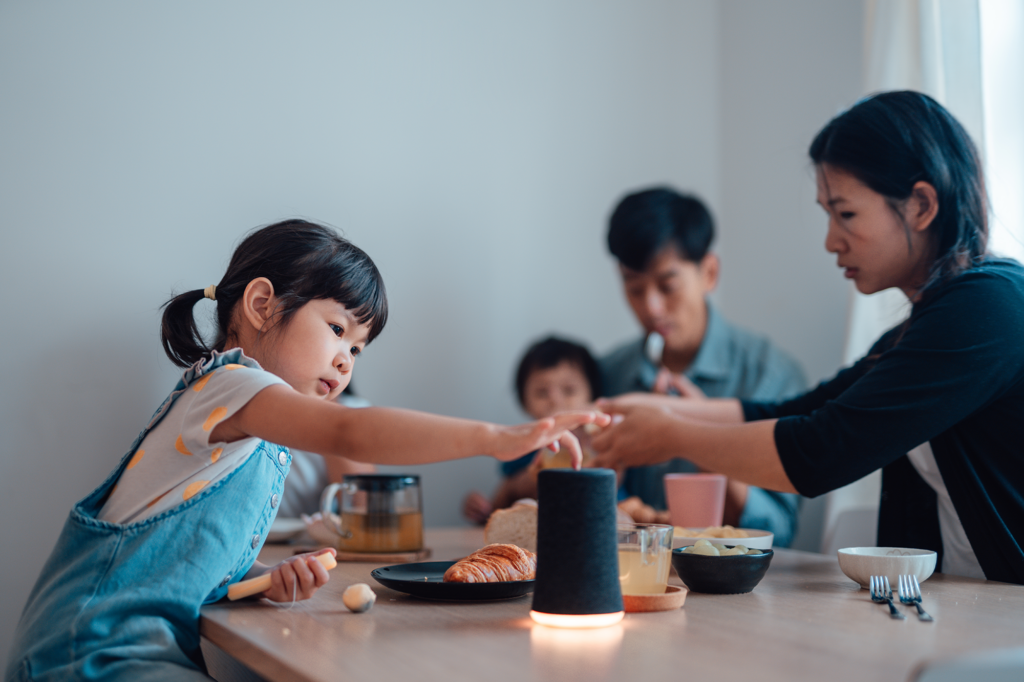 A young girl reaches across a breakfast table with pastries and drinks, while two adults and a child sit together in the background in a softly lit room.