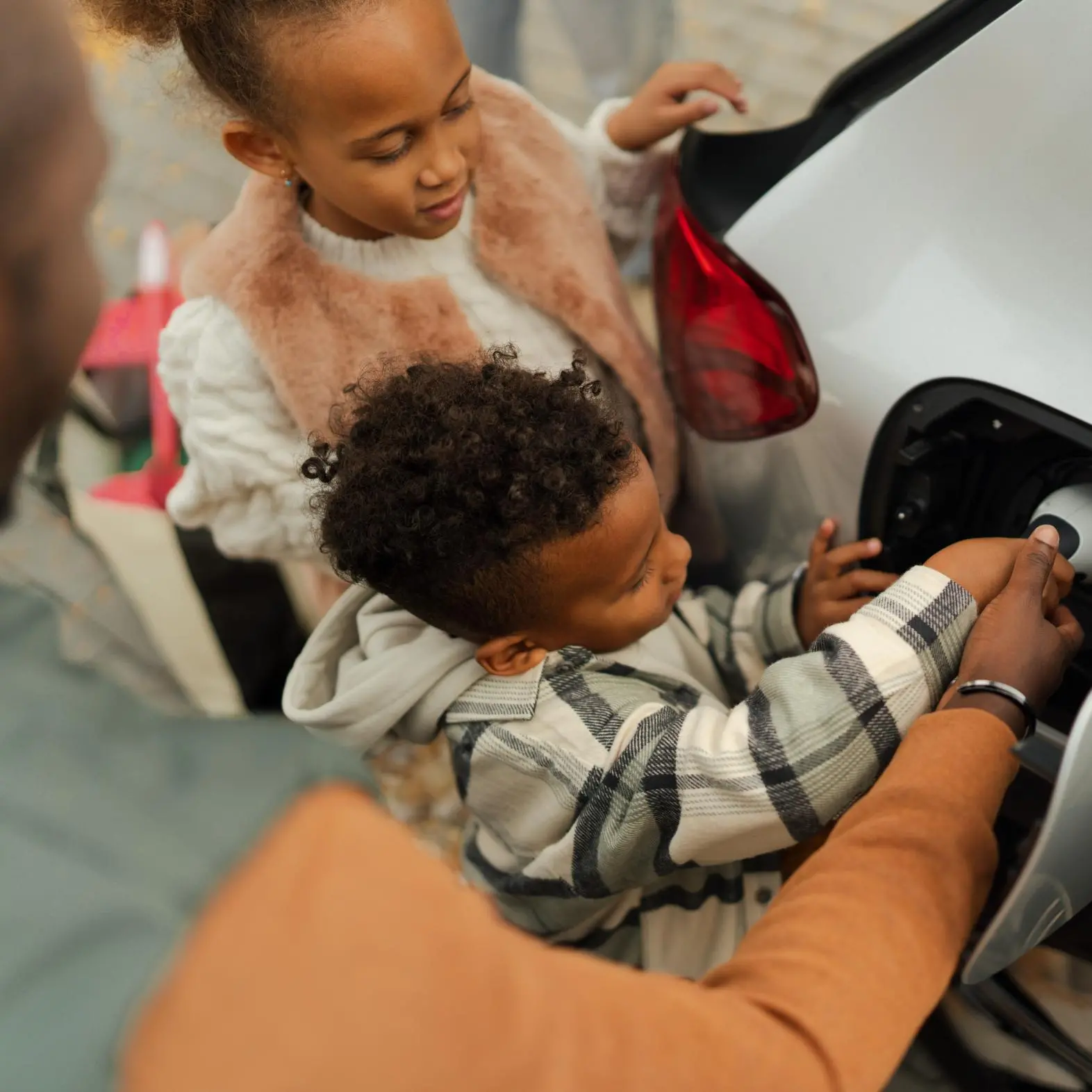 Two children and an adult are standing by an electric car. The child in front is plugging a charging cable into the car while the other child looks on, smiling. The scene appears to be outdoors.