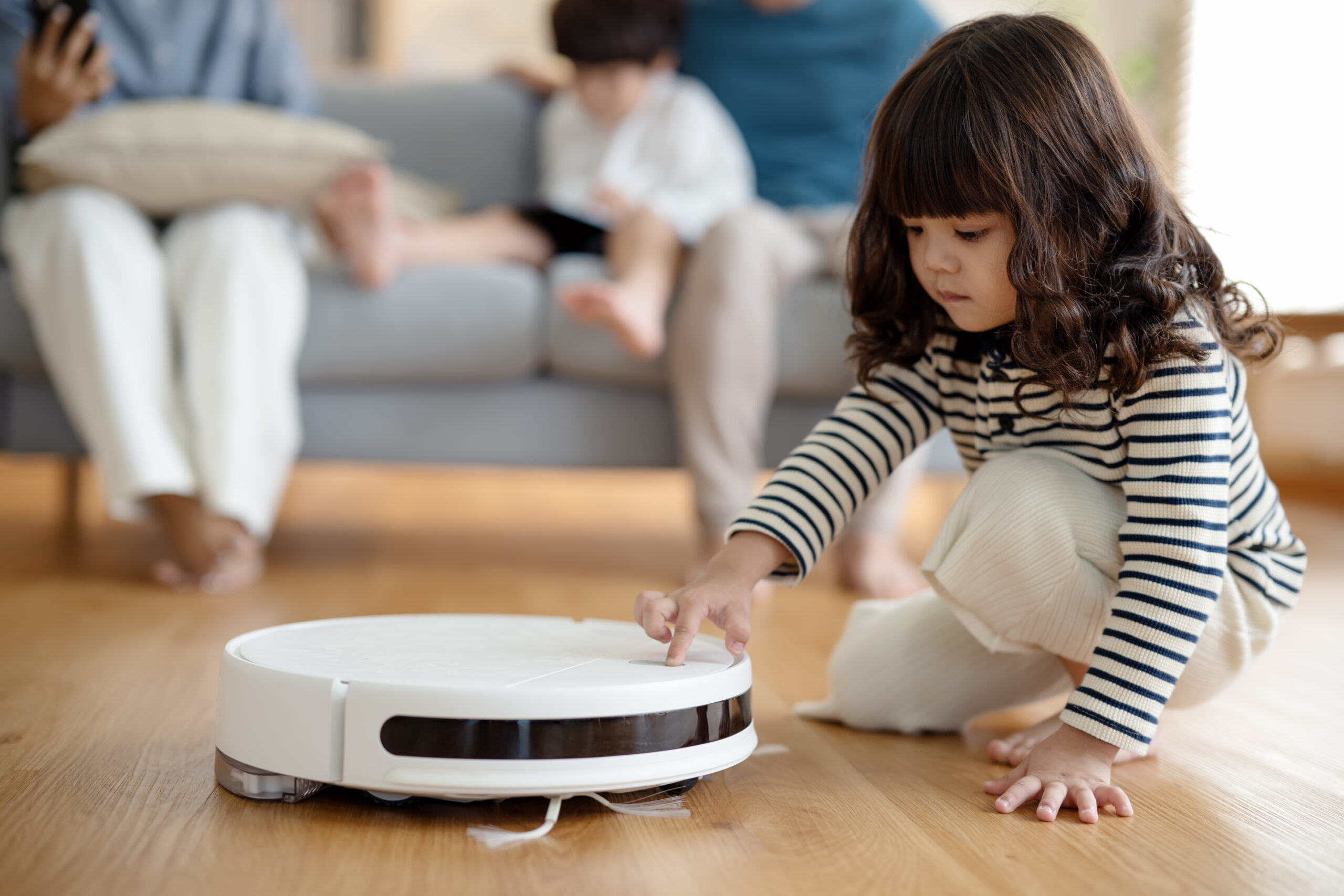 A young girl kneels on a wooden floor, touching a white robotic vacuum cleaner. In the background, two adults and a child sit on a sofa, slightly out of focus.