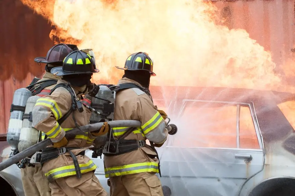 Three firefighters in full protective gear and helmets use a hose to spray water on a burning car, with large flames and smoke rising in the background.
